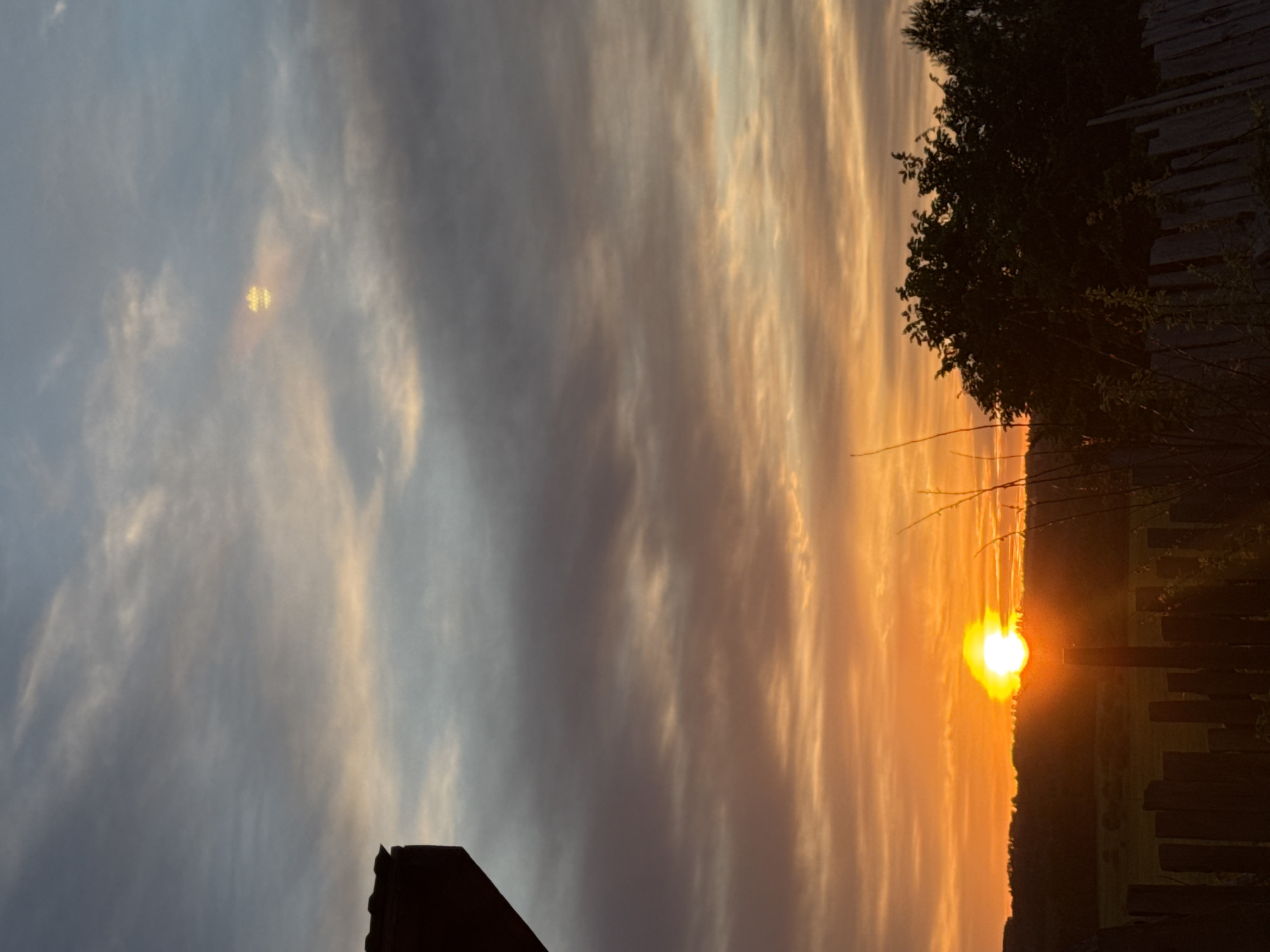 The image depicts a sunset in Nizhnyaya Gar', Russia. The sun is positioned low on the horizon, radiating a bright orange glow. The sky is a mix of grey and orange clouds, with streaks of light filtering through them. In the foreground, there's a silhouetted fence and foliage. The scene appears to be taken from a rural or semi-rural location, with fields visible on the horizon.