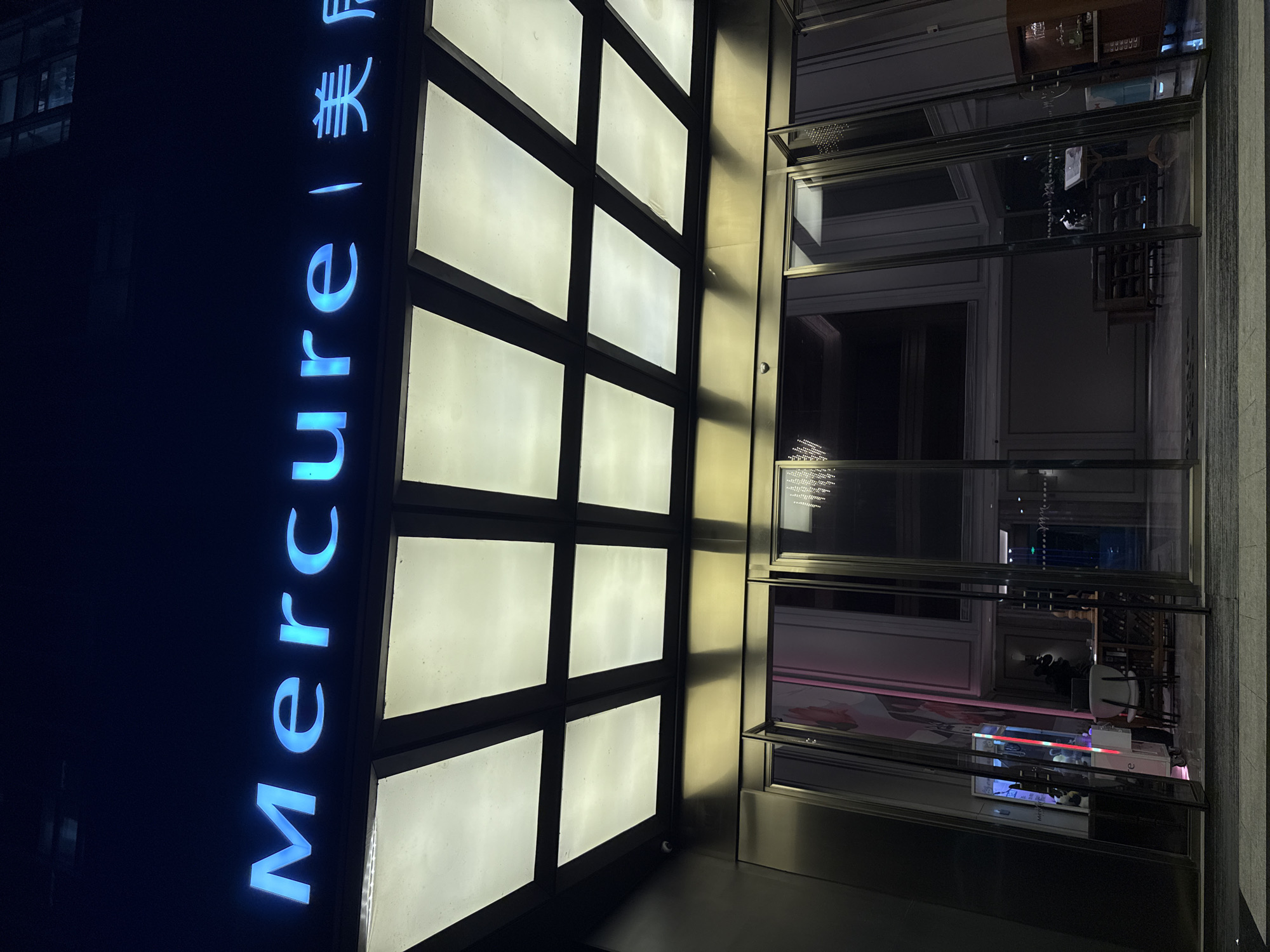 A nighttime, low-angle exterior shot shows the entrance to a Mercure hotel in Huangpu District, China. The hotel's name, "Mercure," is illuminated in bright blue neon letters against a dark background. Above the entrance is a modern canopy with a grid of illuminated panels. The entrance itself features automatic glass doors that are partially open, revealing a glimpse of the hotel's lobby. Inside, elements of the lobby are visible, including decorative lighting fixtures, furniture, and what appears to be a reception area. The overall impression is of a well-lit and contemporary hotel entrance at night.