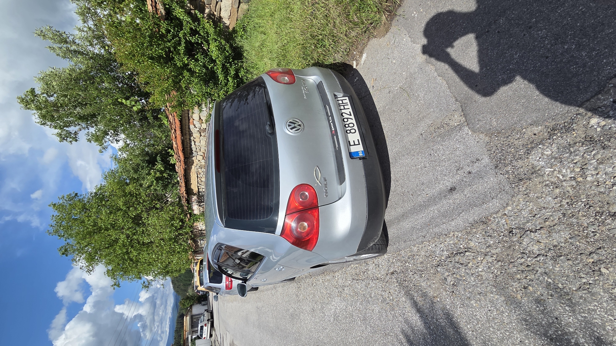 The rotated image captures the rear-right view of a silver-grey Volkswagen Golf (likely a Mk5 model) parked on an asphalt road in Bania, Bulgaria, under clear daylight conditions. The car's tailgate features the circular Volkswagen emblem, "GOLF" badging on the lower left, and "2.0 TDi" on the lower right. A small "sports" decal is located above the license plate, and a silver fish symbol (ichthys) decal is affixed below the "GOLF" lettering.
The vehicle displays a Bulgarian license plate: "E 8892 HH," with the "BG" country identifier on the left. The rear window of the car is tinted. To the left of the vehicle, a stone and brick wall is visible, overgrown with dense green foliage and trees that extend upwards into a bright blue sky dotted with white clouds. In the background, hints of distant mountains and other structures suggest a village or town setting.
The road surface is asphalt, exhibiting signs of wear with cracks and patches of gravel. A prominent shadow of a person, holding what appears to be a camera or phone, is cast on the road in the foreground, indicating the photographer and suggesting a sunny mid-day or afternoon. The scene depicts a serene outdoor environment.