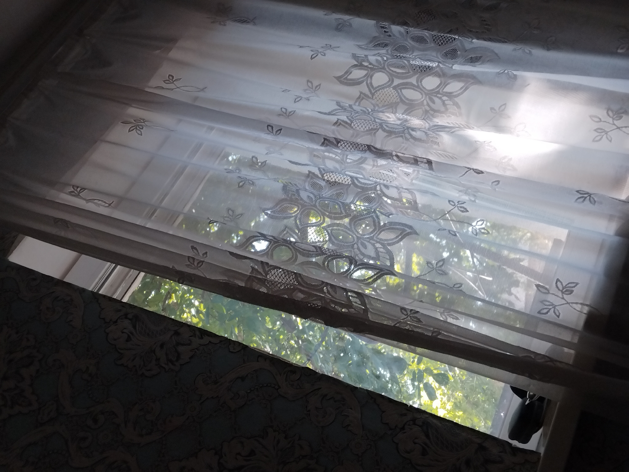 The image shows a window with sheer white curtains with an embroidered floral pattern. Sunlight streams through the window, illuminating the curtains and casting shadows. Outside, green foliage is visible, suggesting a natural setting. The window is framed by patterned wallpaper on the left and a dark wall on the right. A black object, possibly a shoe, rests on the windowsill. The scene appears to be indoors, looking out through a window in Pervozvanivka, Ukraine.