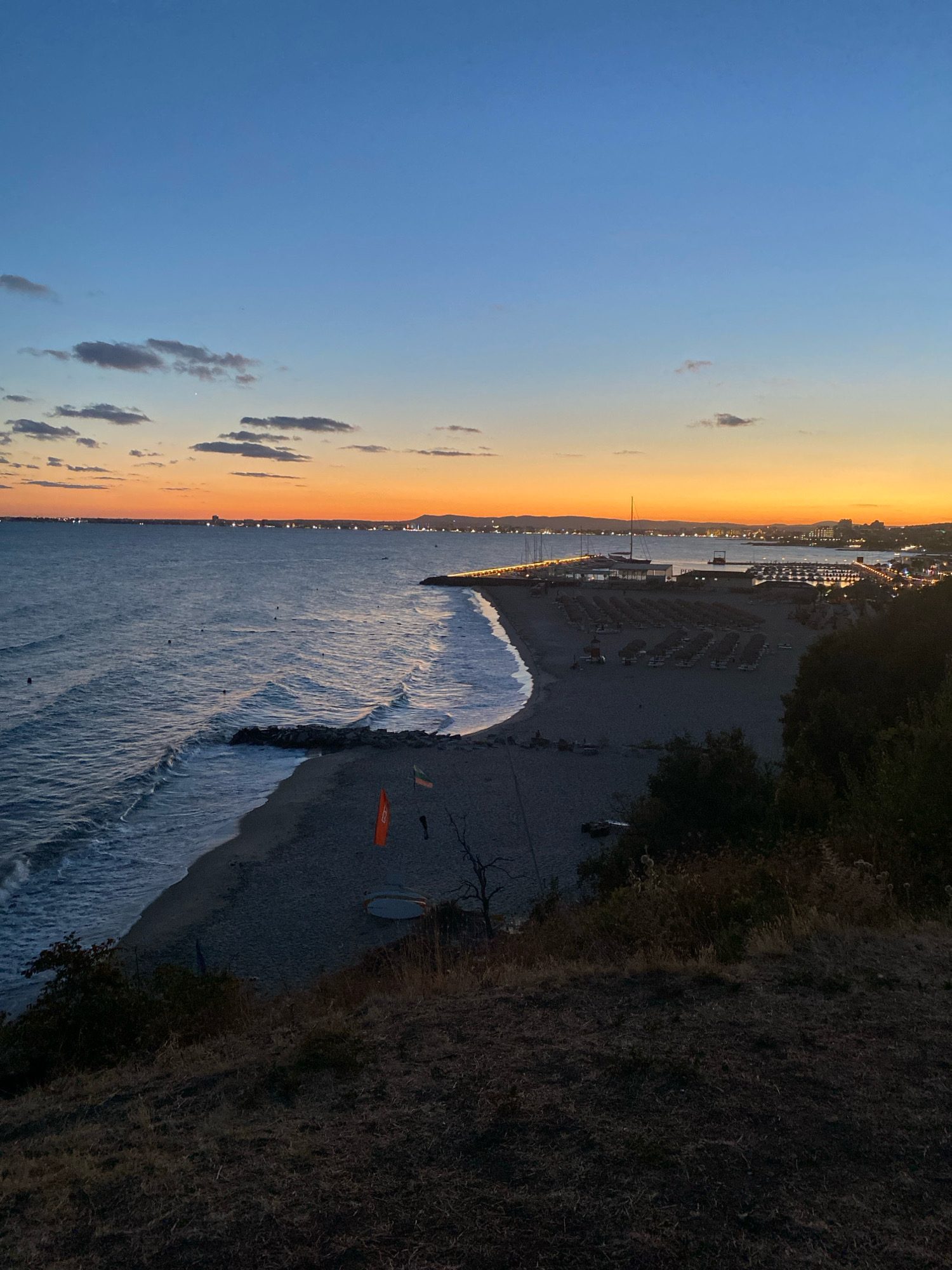 This image captures a serene sunset view over the Black Sea coast in Sveti Vlas, Bulgaria. The sky transitions from a deep blue at the top to a vibrant orange and yellow hue near the horizon, dotted with scattered clouds. The calm sea reflects the fading sunlight, with gentle waves lapping against a pebble beach.
Along the coastline, a pier extends into the water, lined with moored sailboats and yachts. Adjacent to the pier is a beach resort area featuring rows of neatly arranged sun loungers and umbrellas, suggesting a popular tourist destination. In the distance, the lights of Sveti Vlas begin to twinkle as the city embraces the evening. The foreground is dominated by a grassy, elevated viewpoint overlooking the beach and sea, with sparse, dry vegetation characteristic of the Bulgarian coastline. Two orange flags, one triangular and the other rectangular, are planted on the beach, and a white surfboard is visible nearby.