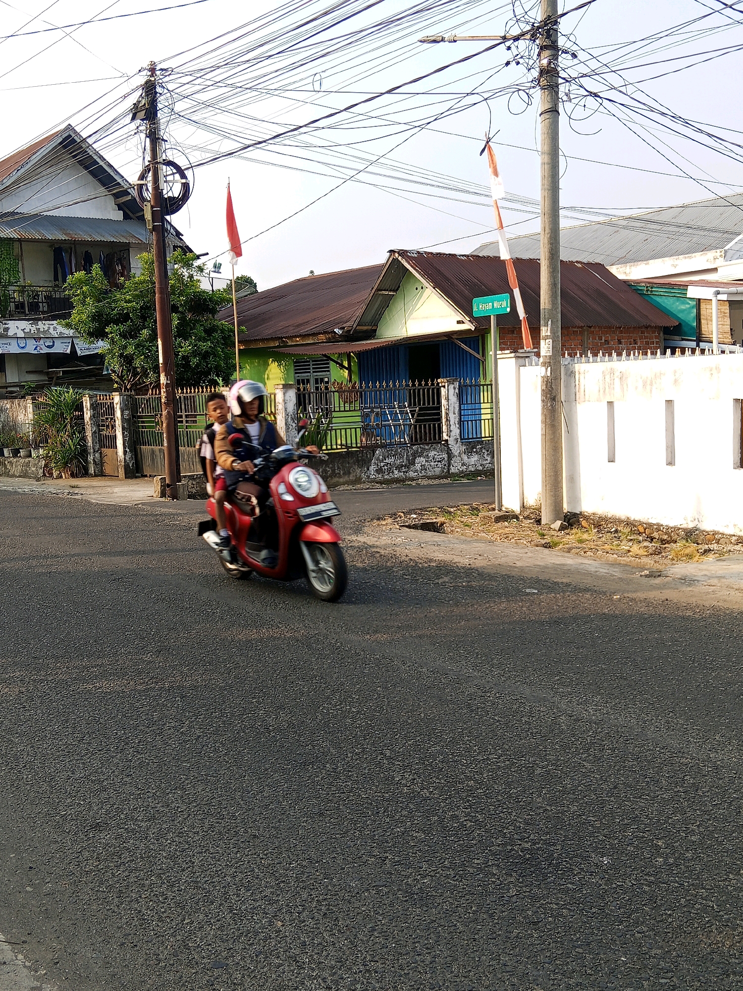 Two individuals are riding on a red scooter on a paved road. The person in the front is wearing a brown jacket and a white helmet, while the person behind is wearing a backpack and a white helmet. The scooter is moving from left to right across the frame. On the right side of the road, there is a white wall with rectangular openings. A utility pole with numerous wires is visible behind the wall. A street sign that reads "Jl. Hayam Wuruk" is attached to the utility pole. In the background, there are houses with varying roof colors, some with visible fences and trees. A red and white Indonesian flag is attached to a bamboo pole, near the street sign and on the left side of the frame, a similar red and white flag is visible on a bamboo pole. The sky is clear and the scene appears to be illuminated by natural sunlight, indicating it is daytime. The overall environment suggests a residential or commercial street in a city.