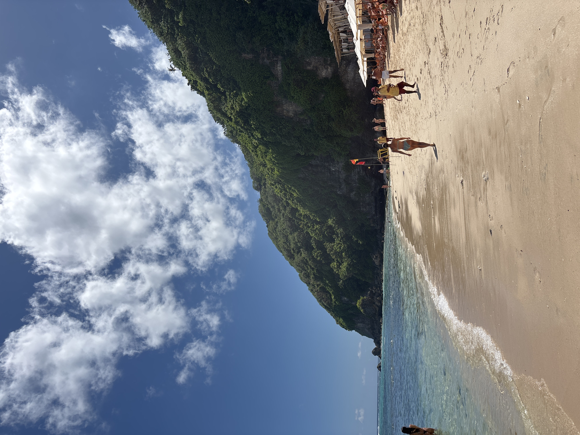 The image shows a beach scene in Kuta Selatan, Indonesia. The beach is sandy with clear blue water lapping at the shore. A steep, green cliff rises along the left side of the beach. People are walking and relaxing on the sand. On the upper edge of the beach, there are structures with people. The sky is blue with scattered white clouds.