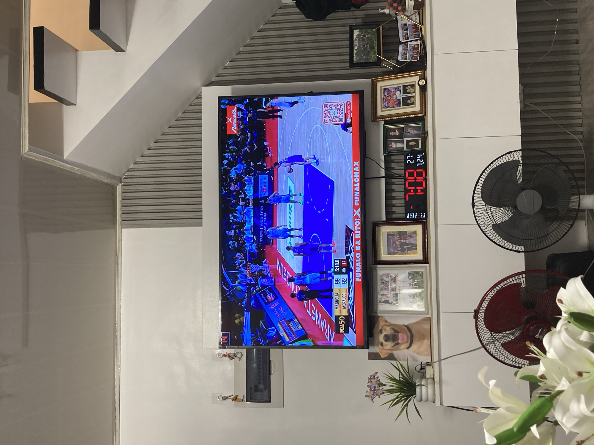 The image depicts an indoor living room scene with a large television displaying a basketball game. The television is mounted on a white wall above a white cabinet. On the cabinet, there are several framed photographs and a digital clock showing "7:08". In the foreground, there are two electric fans, one red and one black, and a bouquet of white flowers. The overall ambiance suggests a casual home setting. The exact location within Davao City, Philippines, cannot be determined from the visual information provided in the image.