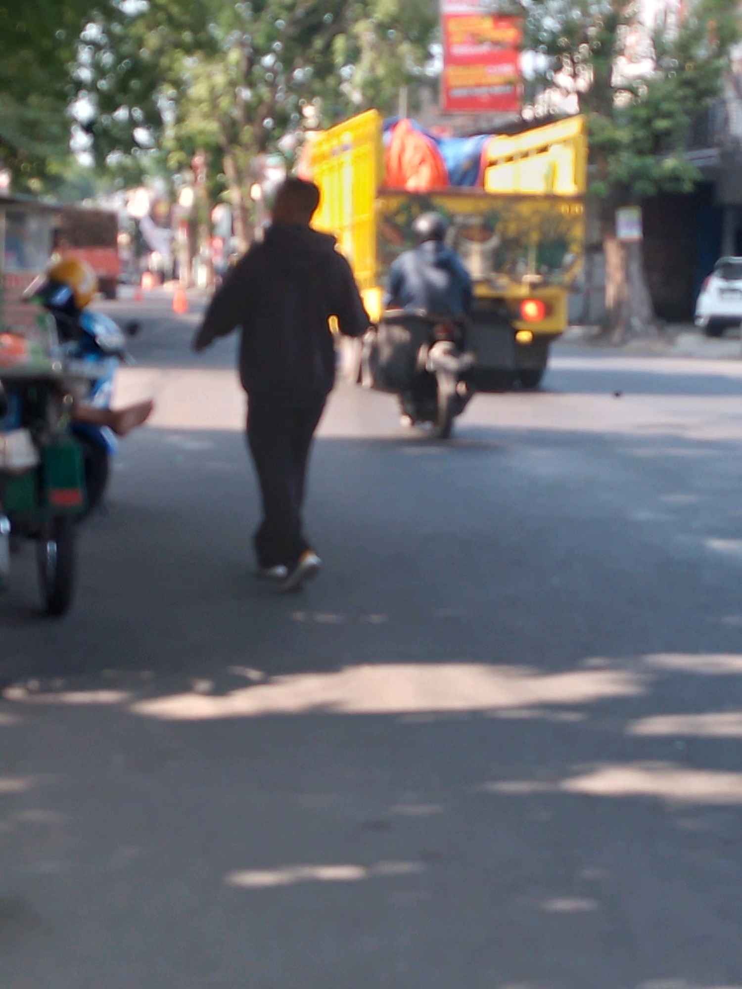 The image captures a street scene in Kertosono, Indonesia, with a focus on several subjects and vehicles. In the foreground, a person is walking away from the camera, wearing a dark hoodie and pants, with their right leg slightly extended. To the left, a motorcycle with a yellow helmet on its seat is partially visible. Further down the road, another person is riding a motorcycle, wearing a dark jacket and helmet. Behind them, a yellow truck with a blue and red tarp covering its load is prominently featured, its brake lights illuminated. The environment appears to be an urban setting with trees lining the street and buildings visible in the background. The lighting suggests it is daytime, with shadows cast on the asphalt. The image has a blurred, low-resolution quality, common in candid street photography or surveillance footage.