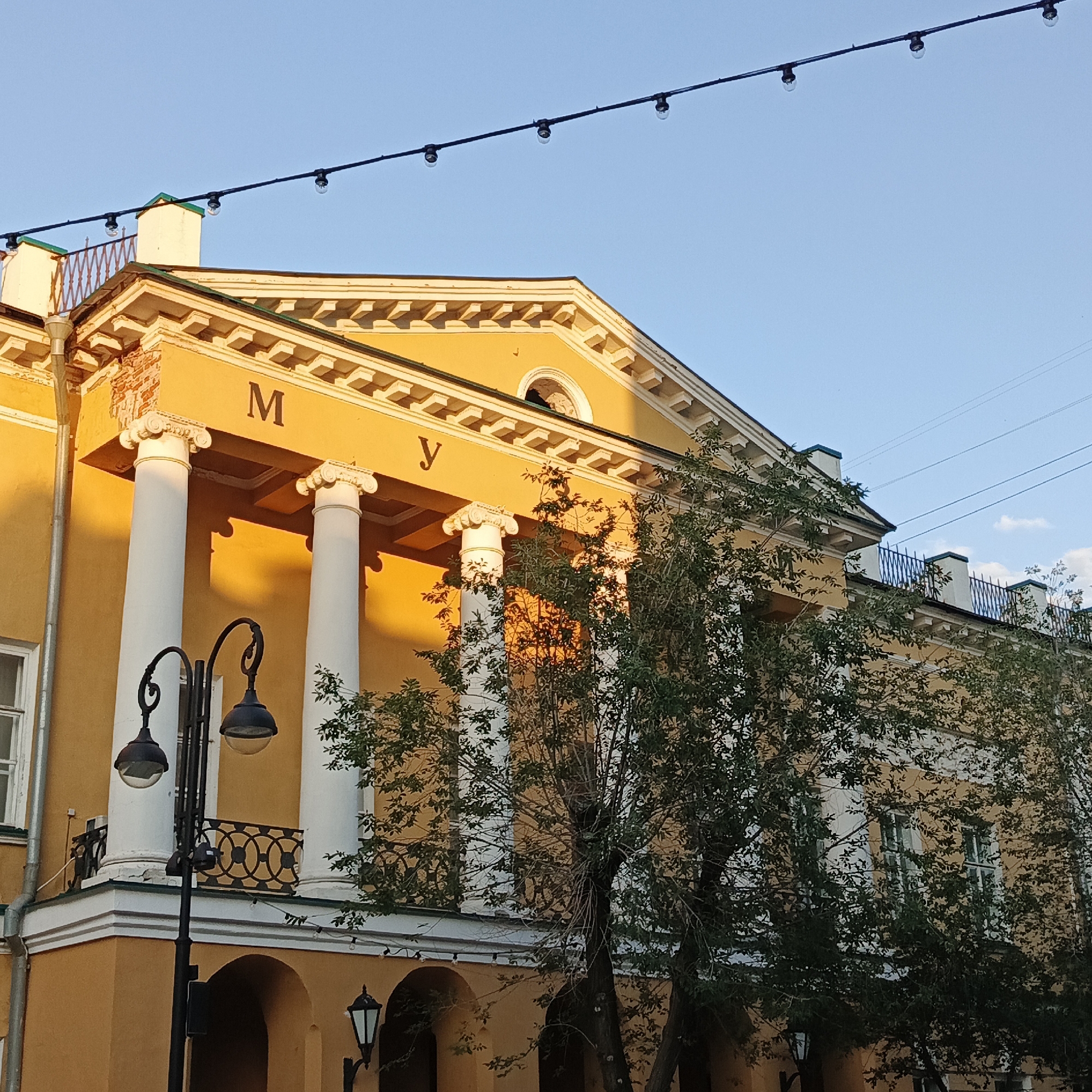 The image shows a close-up of the facade of the Orenburg State Regional Museum of Local Lore. The building is painted a bright yellow and features classical architectural elements, including large white columns with Corinthian capitals. Above the columns, a sign with Cyrillic letters "МУ" is visible. The building has a pediment with decorative molding. A string of lights is strung across the top of the frame, with several bulbs visible. Trees with lush green foliage partially obscure the building. In the foreground, a lamppost with two ornate black lanterns is present. The sky is clear and blue, with some wispy clouds. The lighting suggests it is either early morning or late afternoon, with the sun casting a warm glow on the building.