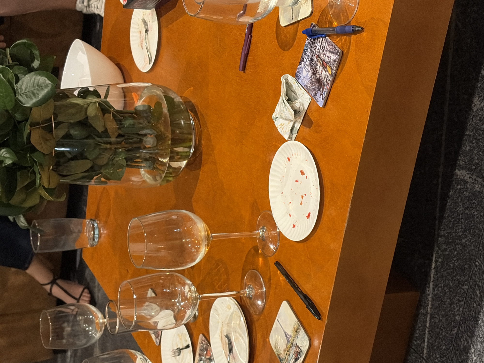 The scene depicts a cluttered wooden table, possibly in a restaurant or event space. There are several wine glasses, some empty and some appearing to be used. Disposable white plates, some with food residue, are scattered across the table along with napkins. A large glass vase containing a bouquet of greenery sits on the edge of the table. Two pens, one purple and one blue, and several artistic coasters depicting possible city scenes are present.
A person's legs and feet, wearing black strappy sandals, are visible near the table's edge.
Given the presence of artwork on coasters potentially indicating a city, it is not possible to confirm the presence of the location of San Bernardino Tlaxcalancingo, Mexico.