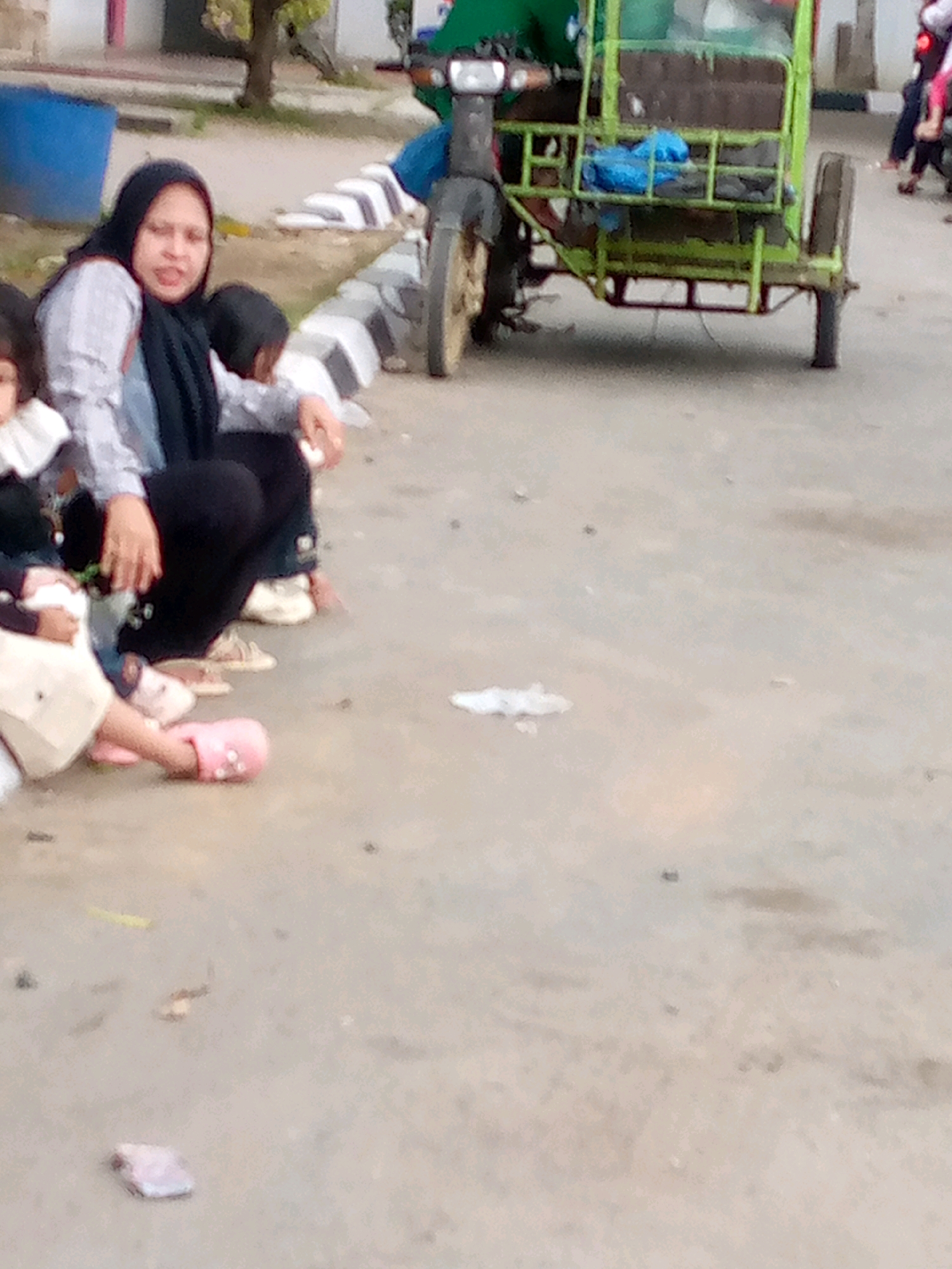 In Keudebayu, Indonesia, a woman wearing a headscarf and a patterned shirt is seated on the ground. Beside her, a child in a white outfit is also seated. In the background, a green tricycle is parked. The ground is paved, and there are some debris and scattered items.