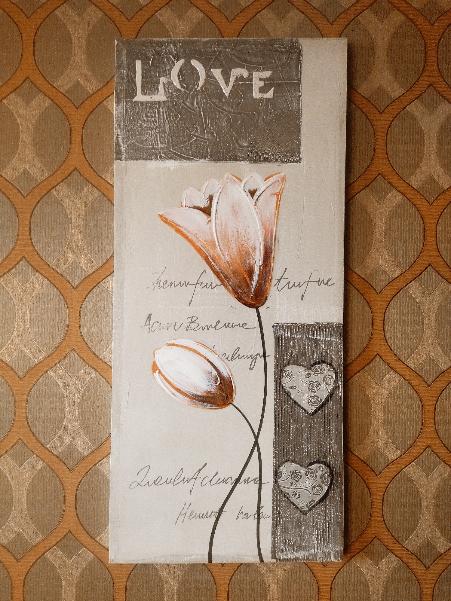 This is a vertical, close-up, eye-level shot of a painting hanging on a patterned wallpapered wall. The painting features a light cream background with a grey textured rectangular area at the top displaying the white text "LOVE" in a stylized font. Below this, two orange and white tulips are depicted. The upper tulip is in bloom, while the lower one is closed. To the right of the tulips, there is a grey textured vertical rectangle with two cut-out heart shapes. Each heart has a floral pattern within. Faint, cursive-like text is scattered across the cream background of the painting, some appearing to be French phrases like "truffe" and "Avoir."
The wallpaper has a repeating pattern of interlinked ovals in shades of gold and light brown, with a textured finish. The lighting is even, suggesting an indoor setting. There are no visible people or animals. The overall mood of the painting appears romantic and serene. The location context is Temse, Belgium.