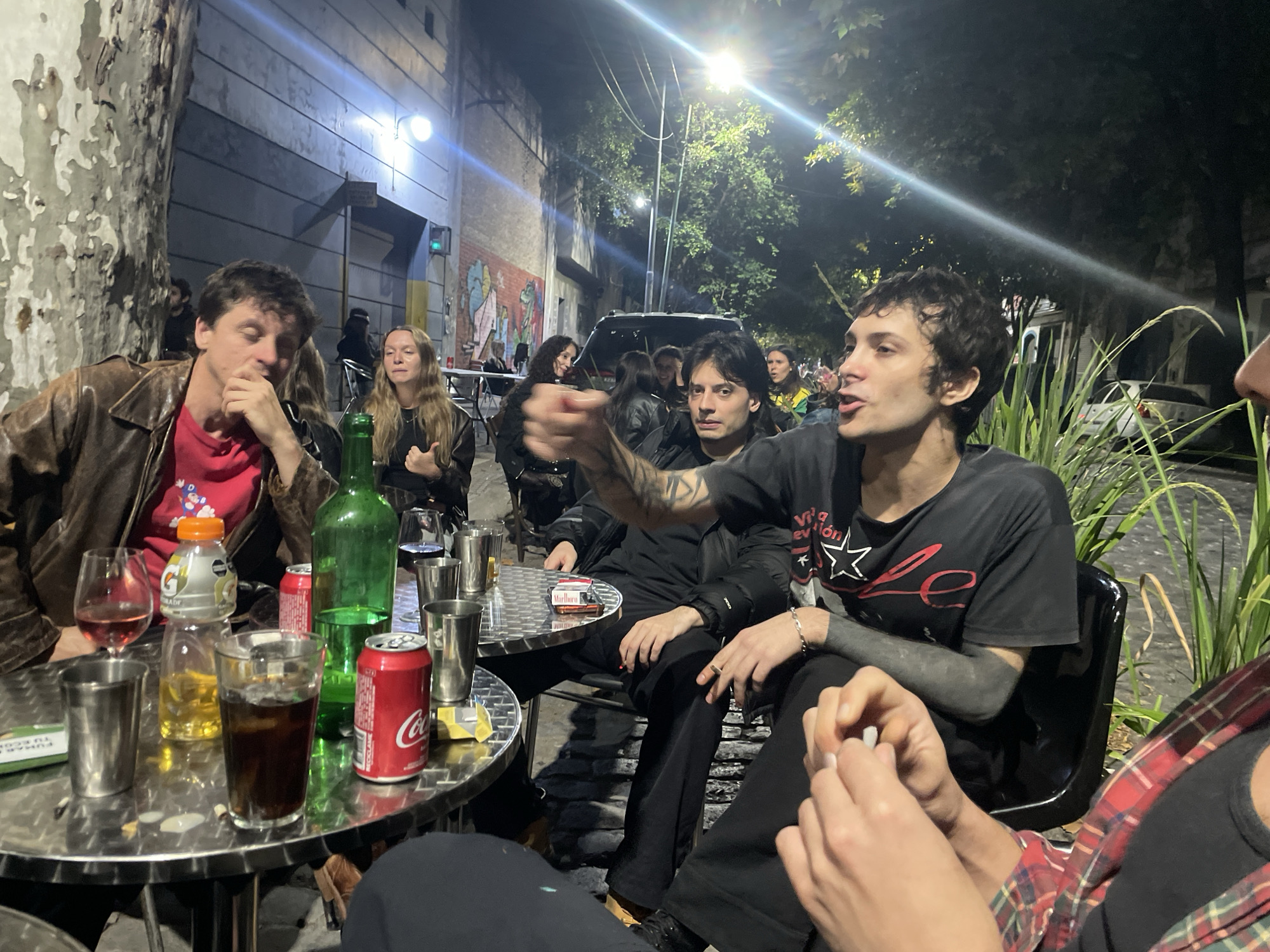 A group of people is sitting at outdoor tables on a street at night. The location is likely a cafe or bar in Buenos Aires, Argentina. The scene is illuminated by streetlights.
People:
- A man in a brown leather jacket and red t-shirt sits at a table, his hand raised to his face.
- A woman with long blonde hair sits at a table in the background.
- Two men sit at another table, one of whom has tattoos on his arm and is pointing.
- Part of another person wearing a red and black plaid shirt is visible.
- Additional people are present in the background.
Objects:
- Tables and chairs are arranged outside.
- Various drinks are on the tables, including wine, beer, soda, and Gatorade.
- Cigarettes and lighters are visible on the tables.
- There are stainless steel cups and a green wine bottle.
- Vegetation grows behind the tables.
- A car is parked on the street.
Actions/Interactions:
- People are sitting and talking.
- One man is pointing.
- Some people are smoking.
Environment:
- The scene is outdoors at night.
- The setting is urban, with buildings and streetlights visible.
- Trees are present in the background.
- There is a mural on a wall in the background.