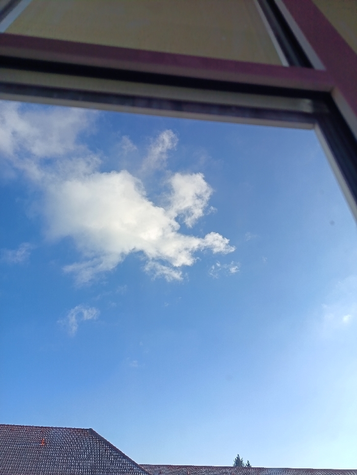 The image presents an outdoor scene viewed through a window frame, visible as blurred dark and reddish-brown structural elements across the top and partially along the right edge. The primary subject is a clear, bright blue sky with multiple white cumulus clouds, notably a larger, more defined cloud formation centered in the upper-left quadrant. The sky on the right side appears brighter, consistent with direct sunlight. In the lower third of the image, the dark grey or reddish-brown tiled roofs of one or more buildings are visible. A small cluster of dark green foliage or trees is observed above the roofline towards the lower-right. No individuals, specific actions, or further identifiable objects are present. This scene is located in the city of Kamenz - Kamjenc, Germany.