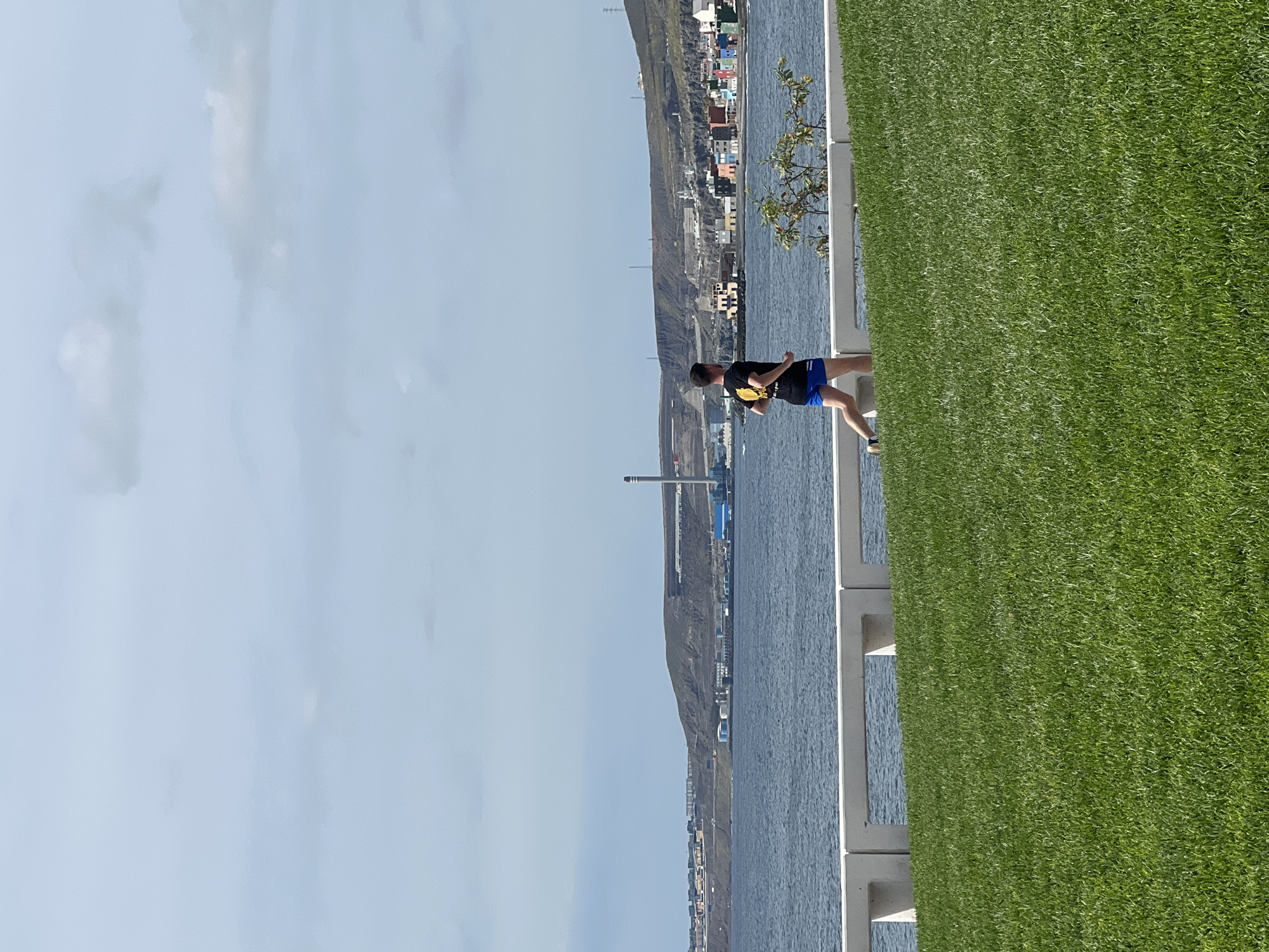 The image captures a scene in Las Palmas, Spain. A man dressed in a black shirt with yellow print and blue shorts stands on a sidewalk, looking out over the water. A grey concrete railing separates the sidewalk from the water, and beyond the water is a rocky, elevated landscape topped with buildings and urban structures. To the man's right is a large patch of green grass or turf. The sky is light blue with some scattered clouds.