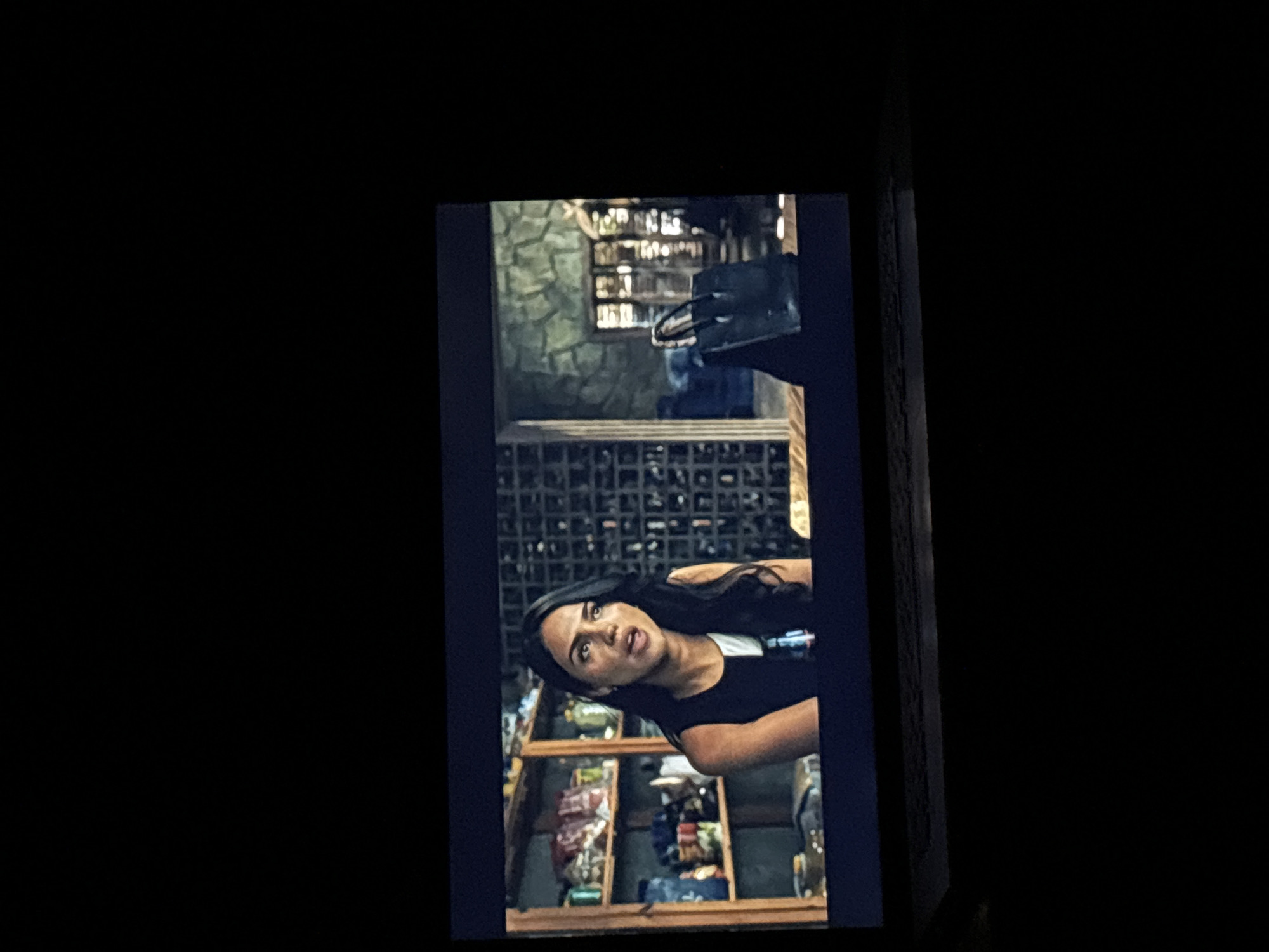 The image displays a digital screen, likely a television or laptop, exhibiting a static frame from a video. The screen is centered within a predominantly dark environment, suggesting a dimly lit room.
The scene on the screen is identified as being located in the city of Budva, Montenegro. It features a woman with long, dark hair, light complexion, and dark eyes, captured from the chest up, positioned slightly to the viewer's left. She wears a sleeveless top that is black on the left side and white on the right side. Her mouth is slightly open, indicative of speaking or reacting.
Behind the woman, the interior setting includes a wooden shelving unit on the far left, stocked with various items such as jars, bottles, and packaged goods. To the woman's right, a wall features a dark, grid-like pattern, possibly a decorative screen or a storage unit. Further right, a section of wall with a rough, stone-like texture is visible. On a surface in the mid-ground to the right, a dark, structured handbag is placed. Beyond the handbag, a window or doorway with vertical mullions allows some light to enter the space. The overall environment on the screen suggests an indoor setting with rustic or textured architectural elements.
The surrounding area outside the screen is uniformly dark, providing no discernible details beyond the screen's illuminated content and a subtle reflection on a surface below the screen, possibly the base of a device.