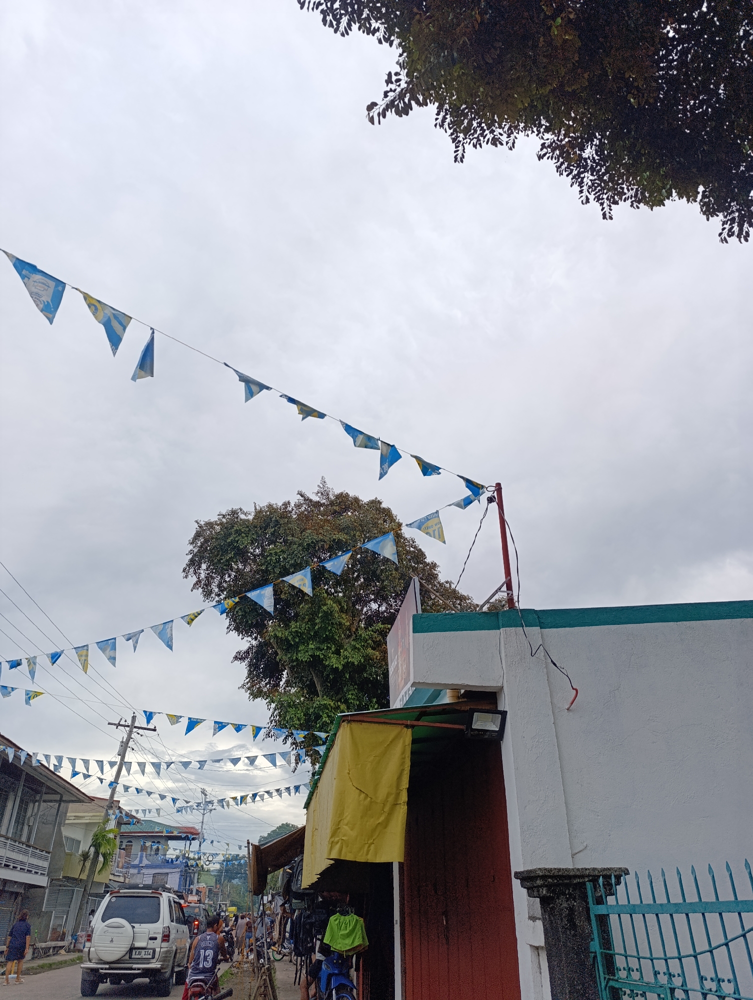 The image captures an outdoor scene in what appears to be a street in a town or city. The sky is overcast and appears to be a cloudy day. A string of blue and yellow triangular flags is strung across the scene, hanging from wires supported by utility poles. These flags are a prominent decorative element.
On the right side of the image, a white building with a teal trim is visible. There is a red-painted wooden door or entranceway on this building, partially obscured by a yellow awning. A black floodlight is mounted above the entrance. A dark, weathered stone pillar stands in front of the building, and a turquoise metal fence is to its right.
On the left side of the image, a street is visible lined with buildings. A white SUV with license plate XJU 334 is parked on the street. In the background, there are more buildings and trees, including a large tree with dense green foliage situated behind the flags. Several people are present on the street, some walking and others on motorcycles. One person is wearing a basketball jersey with the number 13. The overall impression is that of a street scene in a populated area, possibly decorated for an event.
