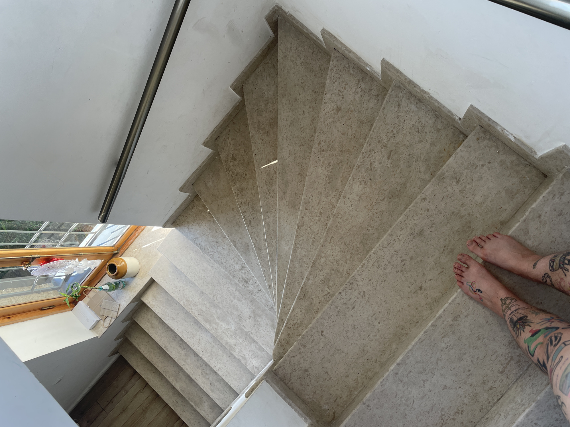This image shows a person's bare feet with extensive tattoos standing on the bottom steps of a winding staircase. The staircase is constructed of light-colored stone with a marbled texture. A white handrail runs along the right side of the staircase. The walls are white. At the top of the stairs, a window with wooden trim overlooks an outdoor scene with greenery. On the windowsill, there are several objects including a white box, two paper bags, a glass bottle with a green plant, and a beige ceramic vase. The floor next to the top of the stairs appears to be wooden planks. The scene is illuminated by natural light streaming in from the window. The overall impression is that of an interior staircase in a residential or public building in Mtskheta, Georgia.