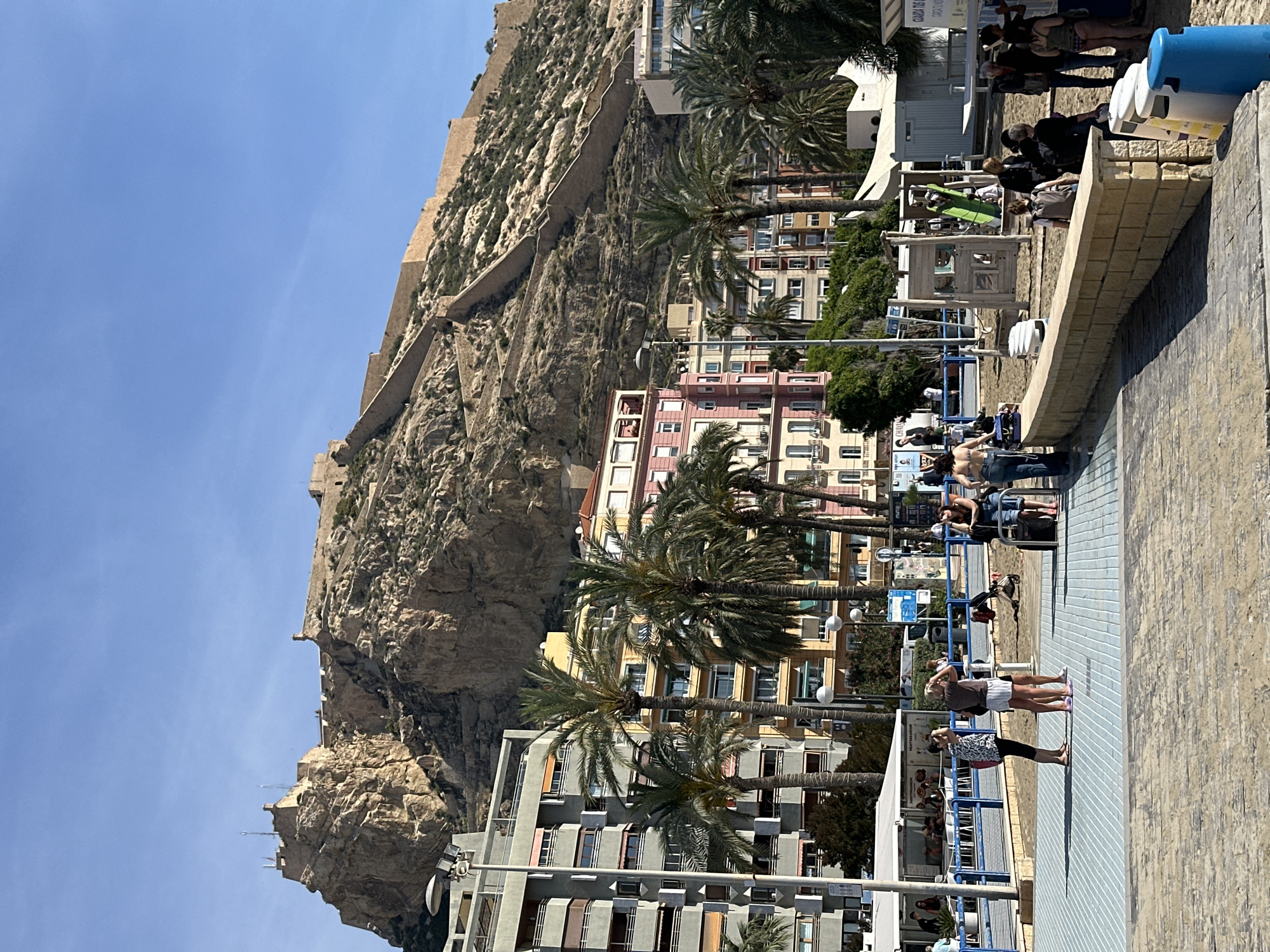 This image captures a scene from Alicante, Spain, featuring Mount Benacantil with the Santa Barbara Castle at its summit. The viewpoint is from a coastal promenade alongside a beach. In the foreground, pedestrians walk along a paved walkway with blue guardrails. Buildings with apartments and palm trees stand behind the pedestrian zone, adjacent to the base of the mountain. Above the buildings, the castle fortifications of Santa Barbara are visible. The sky is clear and blue.