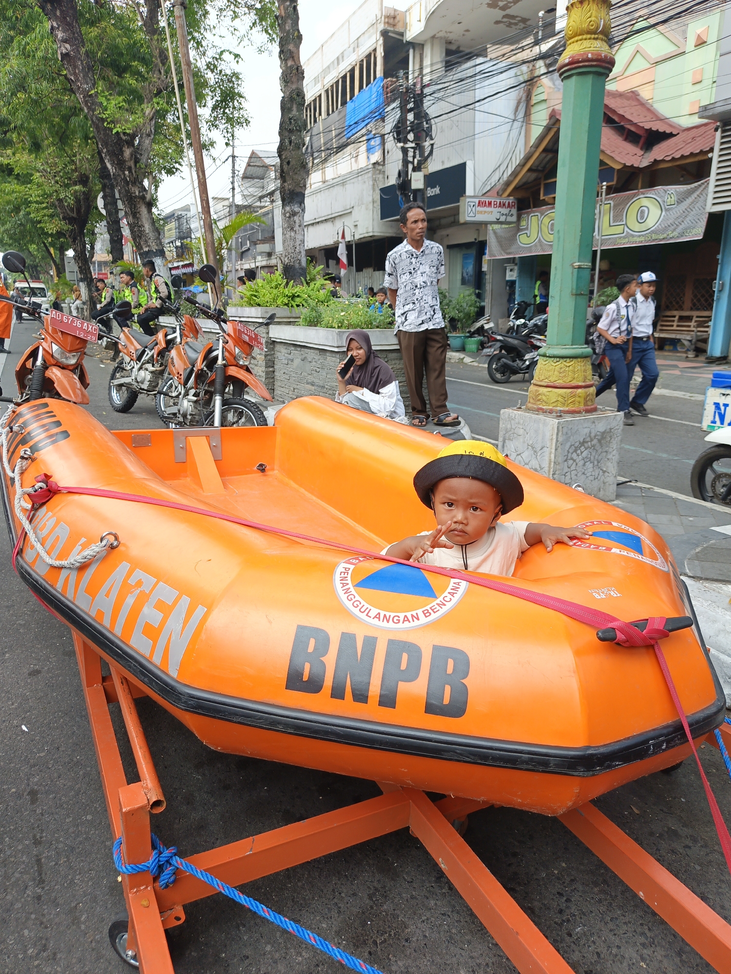 The image depicts an outdoor street scene in Bareng, Indonesia, focusing on an orange inflatable rescue boat on a trailer. A young child wearing a yellow and black hat and a light-colored shirt sits in the boat, looking towards the camera. The boat has lettering on its side, including "KLATEN" and "BNPB," indicating its origin or affiliation. Several orange motorcycles are parked behind the boat, some with people in uniform seated on them. In the background, there are buildings, a tall green lamppost, and more people, including two individuals in school uniforms walking away. The overall impression is of a public space during the daytime, possibly a street or marketplace.