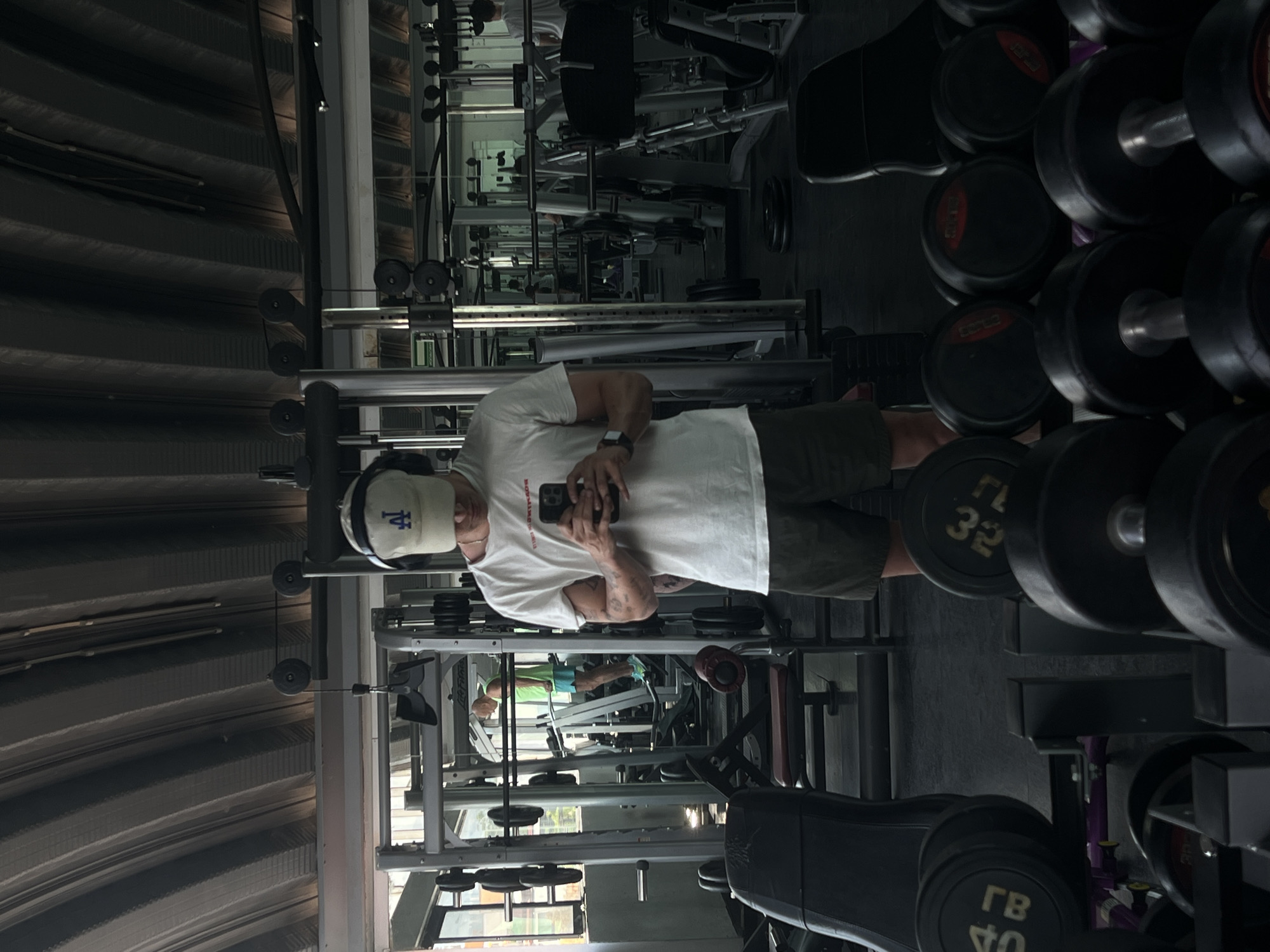 The image is a mirror selfie taken inside a gym in Mexico City, Mexico, depicting a man standing amidst various exercise equipment. The man, positioned in the center-left, is wearing a white t-shirt, dark shorts, and a light-colored baseball cap featuring a blue "LA" logo. He has a smartwatch on his left wrist and is holding a black smartphone with both hands, using it to capture the reflection. His arms show visible muscle definition and tattoos.
The gym environment includes multiple dumbbell racks to the right, filled with black dumbbells of various weights. Visible weight markings on the dumbbells include "2.5 KG," "5 KG," "20 KG," and markings in Cyrillic script such as "ГВ 32" and "ГВ 40." In the background and to the left, an array of weight machines and other exercise equipment is visible. A reflection of another individual, dressed in a green t-shirt and blue shorts, can be seen deeper within the gym.
The ceiling of the gym is constructed from what appears to be corrugated metal. Natural light, likely from a large window in the background, suggests the scene takes place during the daytime. The man appears to be casually posing for the selfie; no specific emotions are clearly discernible.