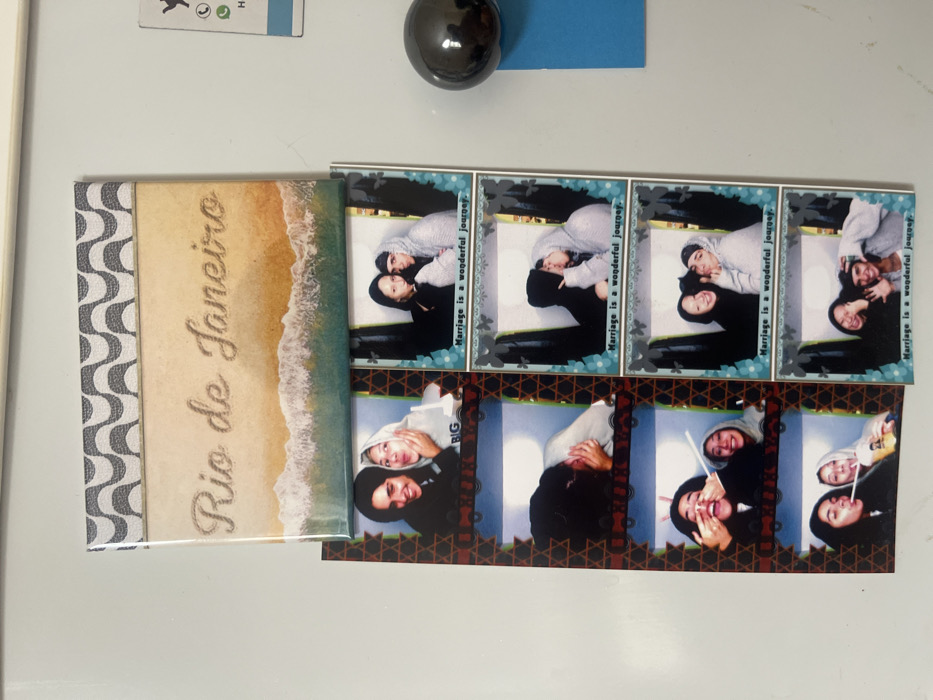 The image displays a white, vertical, possibly metallic, surface, consistent with a refrigerator door. Affixed to this surface are several items.
At the top, a rectangular magnet features a grey and white wavy pattern along its upper edge, resembling the Ipanema boardwalk design. Below this, the magnet depicts a sandy beach scene with "Rio de Janeiro" written in the sand, accompanied by ocean waves.
Centered on the surface are two vertical photo booth-style strips, each containing five individual photographic frames.
The left strip has a reddish-brown border adorned with a repeating star-like pattern. Each frame shows two individuals, likely young women, engaged in playful poses. The person on the left consistently wears a dark top, while the person on the right wears a light-colored hooded sweatshirt. Actions include making faces, holding a cup, drinking with a straw, and making hand gestures.
The right strip features a light blue border with small floral or butterfly designs. Each frame depicts two individuals, one in a dark hooded sweatshirt (partially obscured by shadow in some frames, likely a man) and another in a light-colored hooded sweatshirt (likely a woman). The frames capture intimate and playful interactions, such as kissing, smiling, and making gestures. Each frame on this strip is captioned with "Marriage is a wonderful journey."
To the right of the photo strips, a dark, reflective spherical magnet holds a small, rectangular piece of blue paper against the white surface. Above this, in the top right corner, a portion of a white business card magnet with green and black text/logos, including a WhatsApp icon, is partially visible.
No specific visual cues within the image definitively indicate the location in the city of Buenos Aires, Argentina. The scene appears to be a generic interior domestic setting.