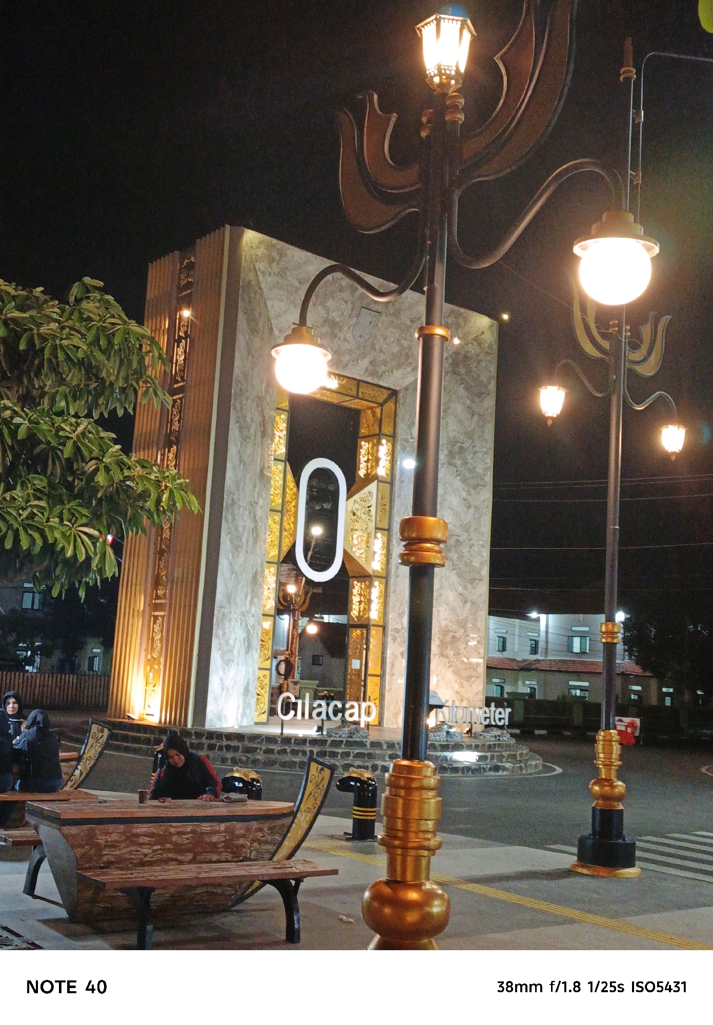 A night-time outdoor scene captures a modern architectural structure with marble-like cladding and decorative gold accents. The building features a large, ornate entrance with a distinctive oval-shaped illuminated sign. In front of the structure, several distinctive, boat-shaped benches with wooden seating are arranged. Two women, one in a black hijab and the other in a black hijab with a red jacket, are seated at one of the benches. A cup sits on the table portion of the bench. Ornate streetlights with golden decorative elements and glowing lamps are positioned throughout the scene. The text "Cilacap" is visible on the architectural structure, identifying the location as Cilacap, Indonesia. The background shows buildings and a street, with evidence of street lighting and a pedestrian crossing. The overall ambiance is illuminated and somewhat festive.