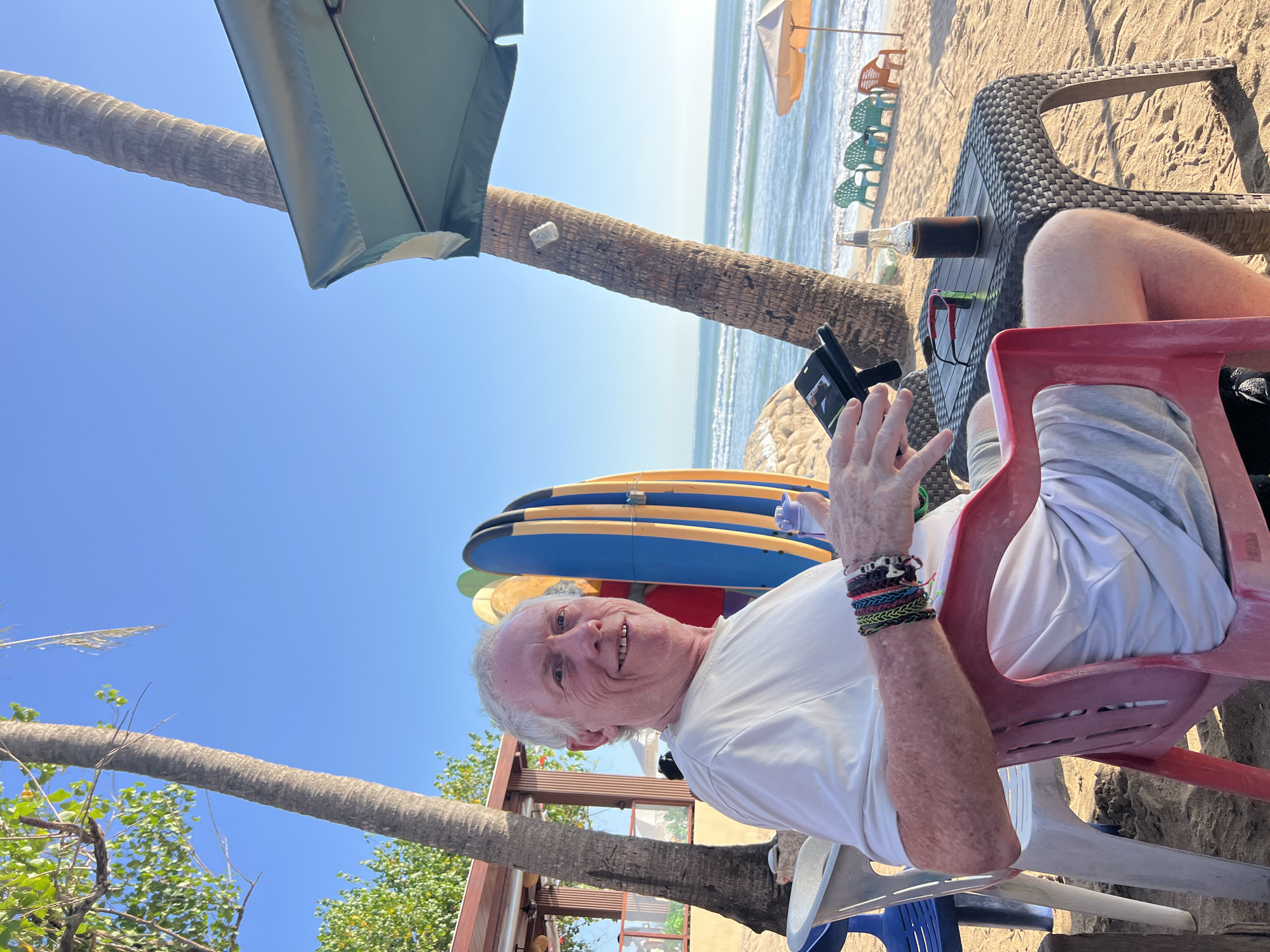 A white-haired man is sitting in a red plastic chair on a beach in Kuta, Indonesia. He is wearing a white t-shirt and light-colored shorts. He has several bracelets on his left wrist. In his hands, he is holding a smartphone and a bottle of what appears to be a dark beverage, possibly beer, in a koozie. In front of him is a black woven table, on which also rests a pair of sunglasses and the beverage bottle. Behind him, several surfboards are propped upright in the sand, and a tall palm tree is visible. The ocean with moderate waves is in the background, and beach chairs and umbrellas are scattered further down the shore. The sky is clear and blue.