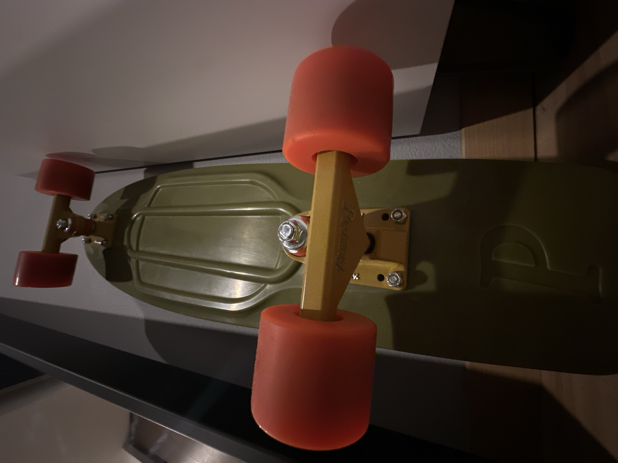 A low-angle, close-up shot shows a green Penny board leaning against a white wall and a wooden shelf. The board has orange wheels and yellow trucks. The background is blurred.