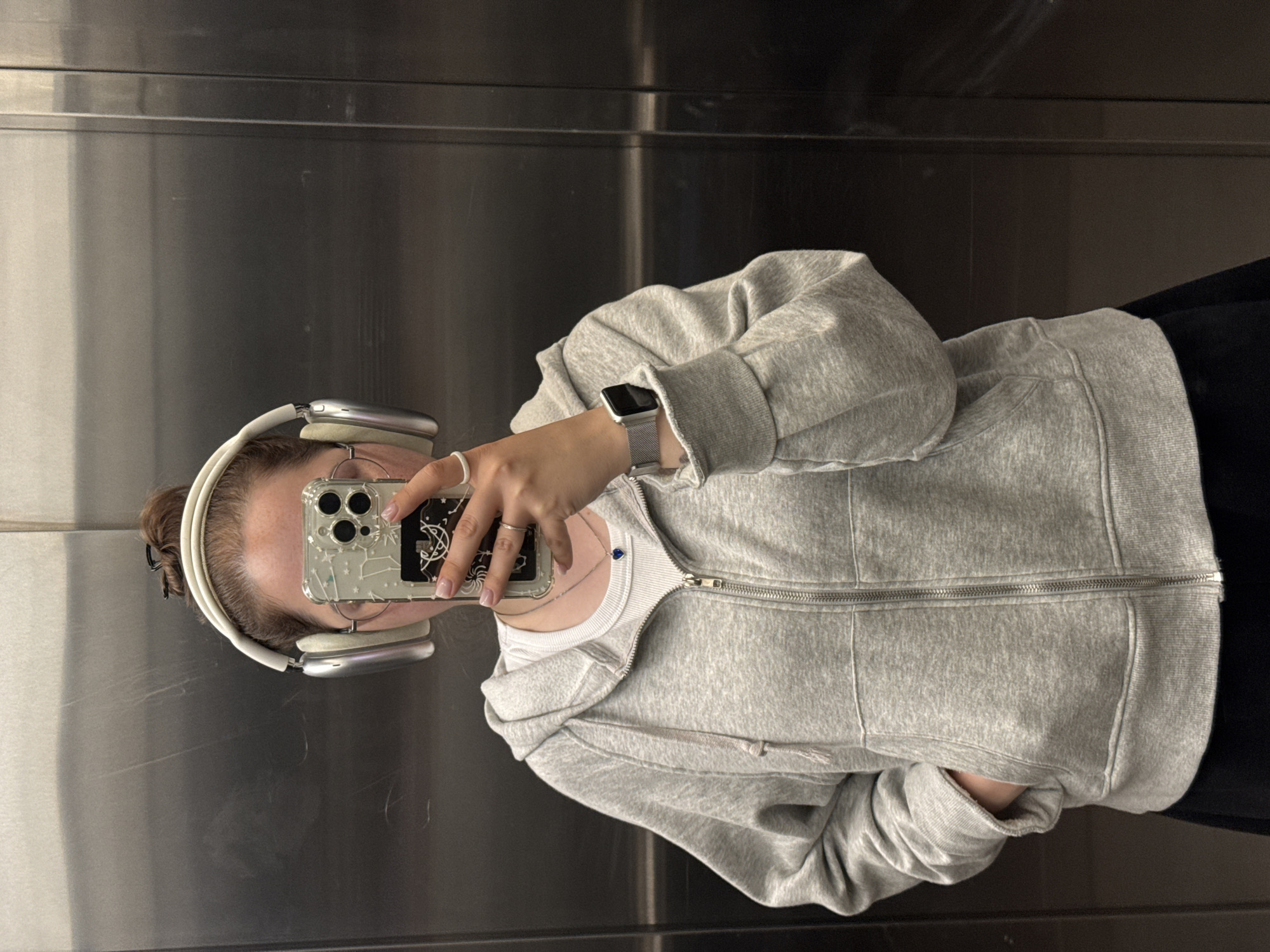 A person is standing in front of a metallic wall, possibly in an elevator, holding up a phone to take a selfie. They are wearing large white headphones, a grey zip-up hoodie over a white tank top, and a silver watch. The person has their hair in a bun and is wearing a necklace with a blue heart pendant and rings on their fingers. The background is a brushed metallic surface with visible vertical lines, suggesting it might be an elevator door or wall. The lighting is even, and the scene appears to be indoors. The location is not specified in the image or its context.