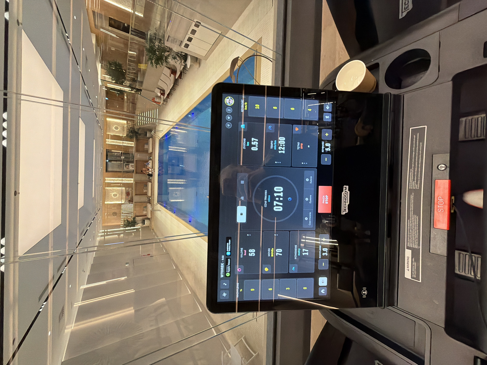 Here is a forensic description of the image:
**Scene:** The image depicts a close-up view of a treadmill console, seemingly in a gym or fitness center overlooking a luxurious indoor swimming pool. The scene is well-lit and modern.
**Objects:**
* **Treadmill:** A black treadmill with a prominent console showing various workout metrics. The console displays data such as speed (5.0 km/h), distance (0.57 km), time (12:00), heart rate, calories burned (58 kcal), incline (3.0%), and elevation (17 meters). Buttons for 'STOP' are visible on the console. There is also a cup holder with a paper cup in it.
* **Swimming Pool:** A large indoor swimming pool is visible through the glass barriers in the background.
* **Gym Interior:** The background shows a well-designed gym interior with lounge chairs, plants, and other gym equipment.
**Environment:**
* **Light:** Bright, artificial lighting illuminates the scene.
* **Modern Architecture:** The gym appears to be located in a modern building with clean lines and a spacious layout.
**People:** No people are visible in the foreground of the image, but there may be individuals in the background near the pool.
**Actions:** The treadmill is in use, based on the metrics displayed on the console.
**Interactions:** The person operating the treadmill is likely interacting with the console, adjusting the settings as needed.
**Additional Details:**
* The console is displaying metrics on Tuesday at 11:32.
* A treadmill brand name "TECHNOGYM" is visible on the console.
In summary, the image shows a treadmill workout in progress at a gym overlooking an indoor swimming pool, characterized by a modern and luxurious environment.
