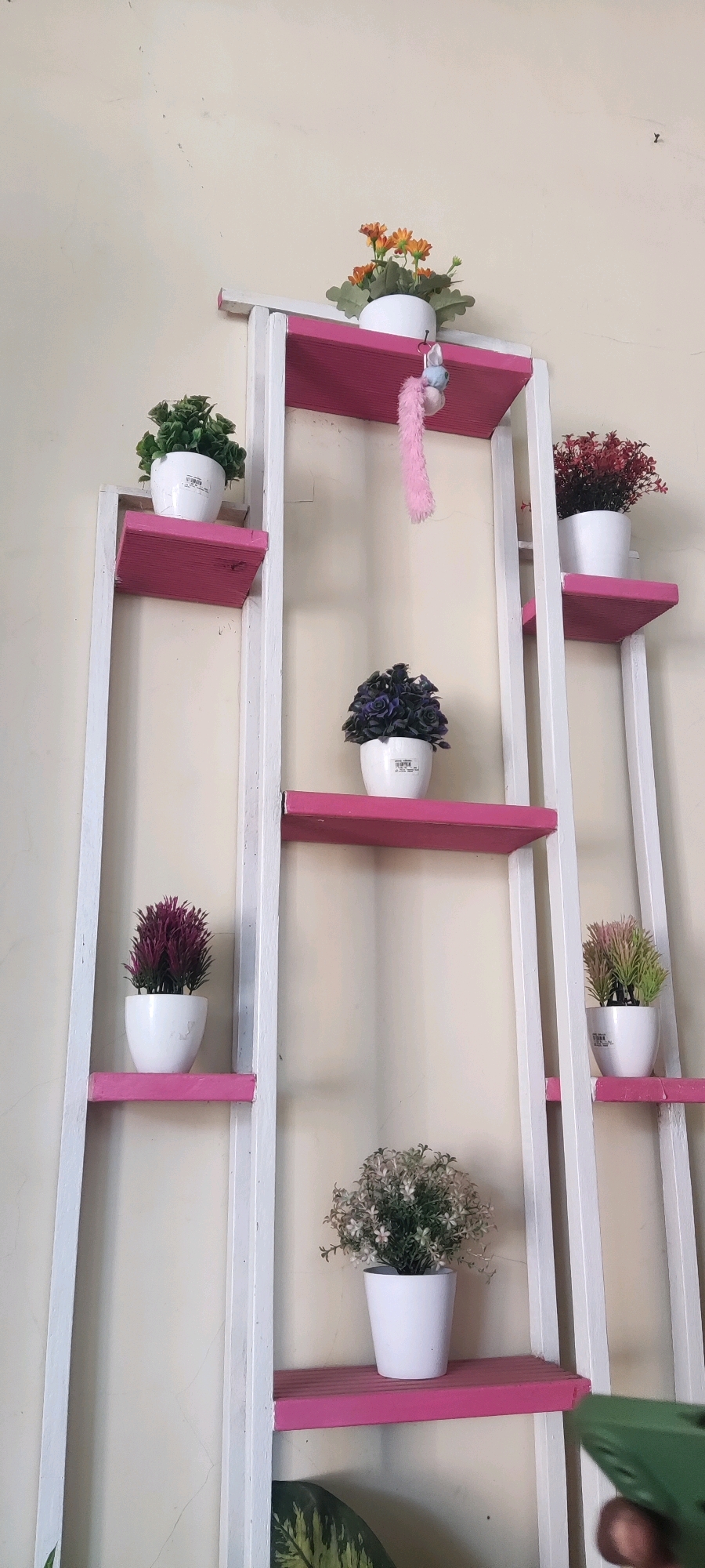 The image depicts a white and pink tiered shelving unit filled with various potted artificial plants. The shelving unit is positioned against a plain, light-colored wall. On the top shelf, there is a small arrangement of orange flowers. Moving down, the shelves hold plants with green foliage, a dark purple plant, a reddish-purple plant, and a plant with green and pinkish-green leaves. The bottom shelf, partially visible, holds a plant with small white flowers and green leaves. A green object, possibly a remote control or a phone, is held by a hand in the bottom right corner, partially obscuring the lower part of the shelving unit and the wall. The lighting suggests an indoor environment, likely a home. There is no specific indication of Japura Kidul, Indonesia, in the visual elements of the image itself.