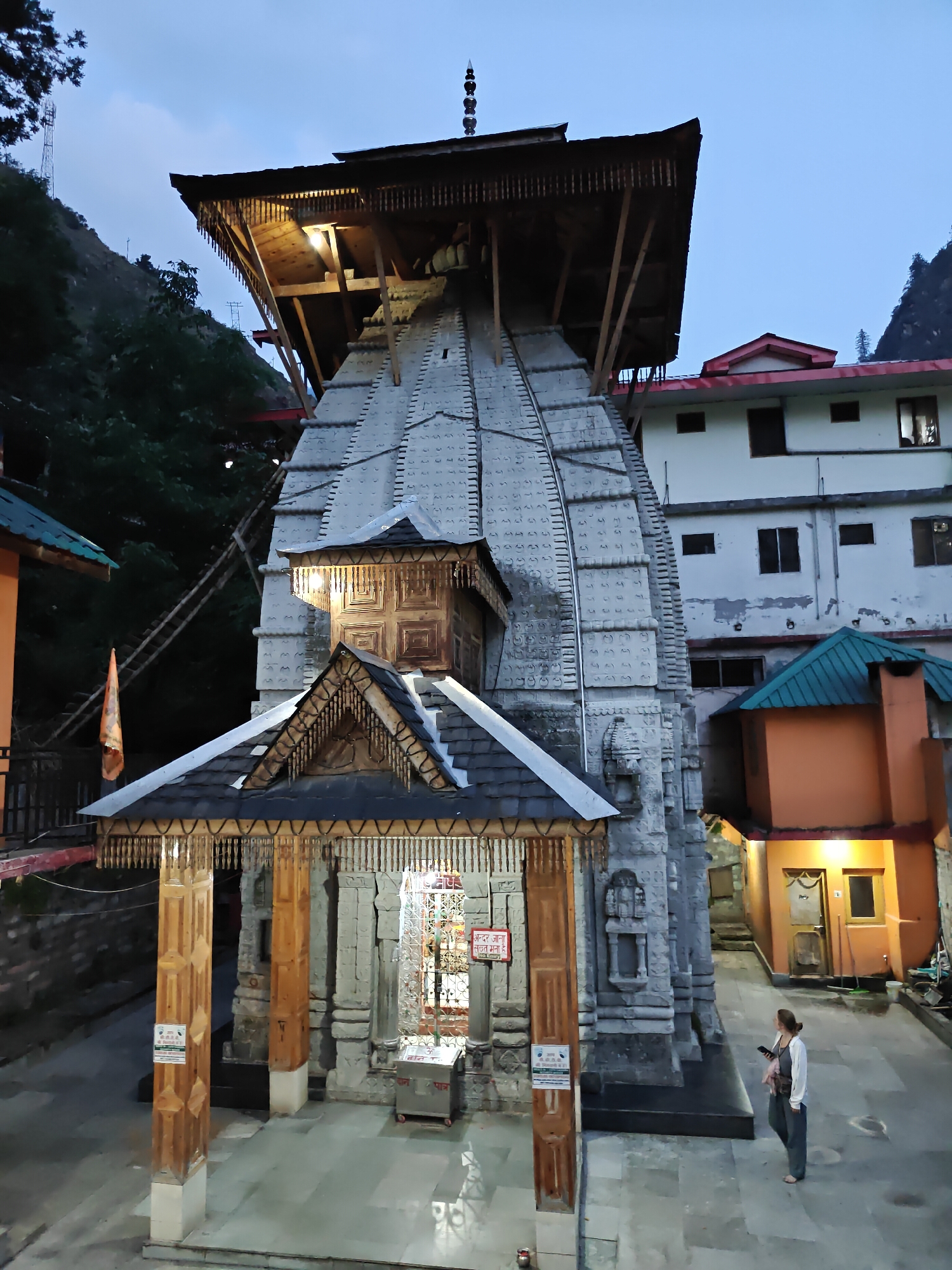 An evening view captures the ancient Shree Raghunath Temple in Manikaran, India, amidst mountainous terrain. The central subject is a multi-tiered stone temple, characterized by a grey, conical shikhara (spire) adorned with intricate carvings and vertical bands. The temple features a prominent, multi-level wooden roof structure, supported by wooden beams and featuring ornate eaves with decorative wooden elements. A distinctive pinnacle crowns the very top.
Artificial lights illuminate the temple's wooden elements and main entrance, casting a warm glow. The temple's facade also includes a smaller, ornate wooden structure above the main entrance, which itself is sheltered by a sloped wooden roof supported by tall, carved wooden pillars.
To the right of the temple stands a large, multi-story building with white walls and red-tiled roof sections, appearing to be a modern structure. Smaller, colorful buildings with orange walls and green roofs are also visible nearby, blending with the temple's surroundings. The background shows steep, tree-covered hills under a twilight sky, indicating either dusk or early morning.
In the lower right, a woman dressed in a white shirt and dark trousers stands looking at her phone, observing the scene. The ground is paved, suggesting a courtyard or open area in front of the temple.
Visible text includes a red sign near the temple entrance stating "अन्दर आना सख्त मना है" (Entry strictly prohibited) and a metal donation box with "ॐ दान पात्र" (Om Donation Box) written on it. Another white sign below the red one is present, but its text is illegible. The overall atmosphere is serene and devotional, highlighting the temple's architecture and its mountain setting.