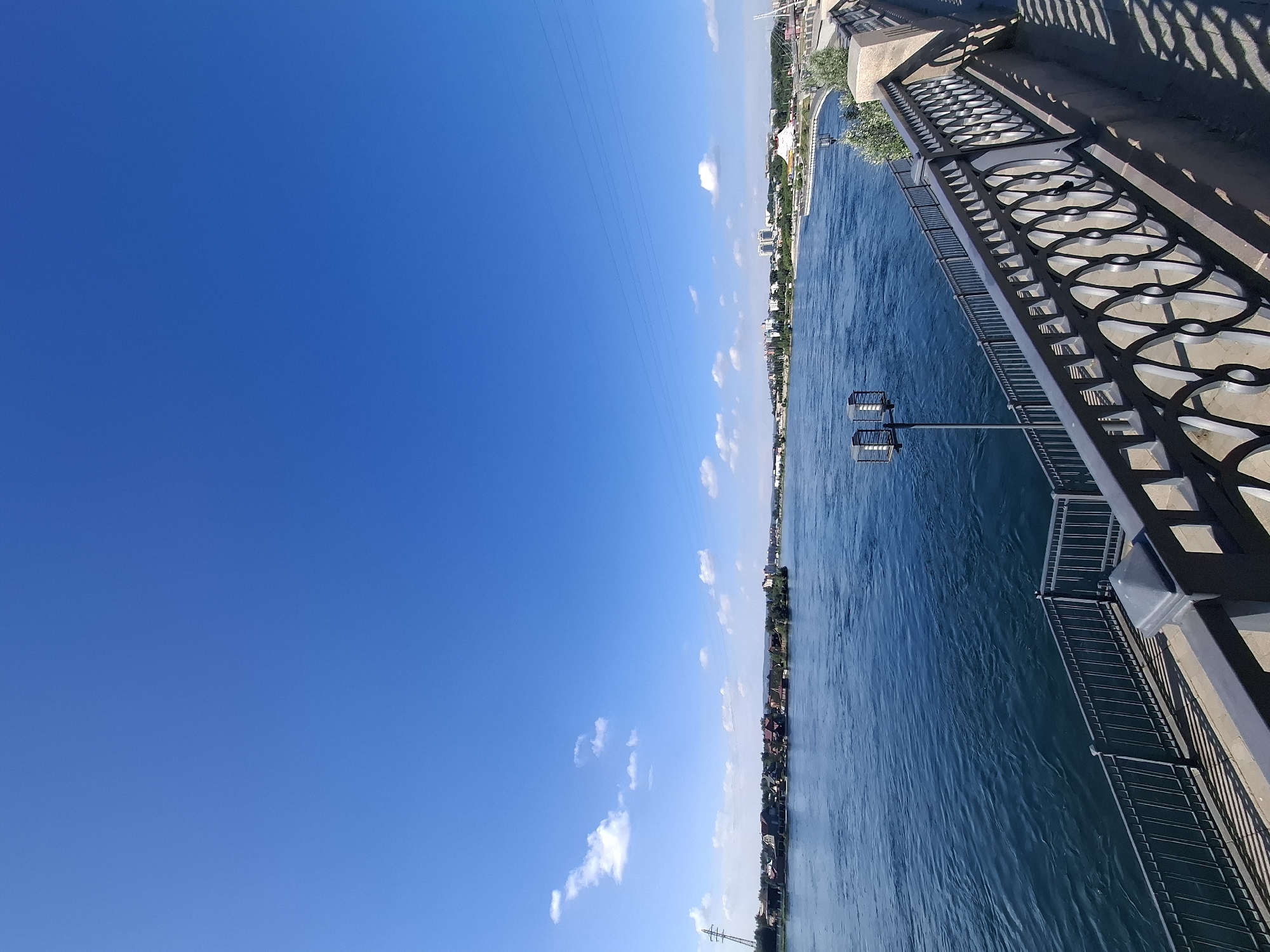 This is a photograph of a wide river in a city. The river is deep blue and appears to be flowing. The sky is clear and bright blue, with a few white clouds scattered across it. On the far bank of the river, there is a line of houses and trees, and in the distance, buildings can be seen, suggesting a city skyline. In the foreground, there is a paved walkway with a decorative metal railing. A lamppost is visible on the walkway. The scene appears to be a waterfront promenade.
Based on the visual elements and information available about Irkutsk, Russia, this scene is likely the Angara River embankment in Irkutsk. The architecture of the buildings on the far bank, as well as the style of the promenade and railing, are consistent with known public spaces in the city. The presence of the wide river and the city skyline are characteristic of Irkutsk's geographical features and urban landscape.