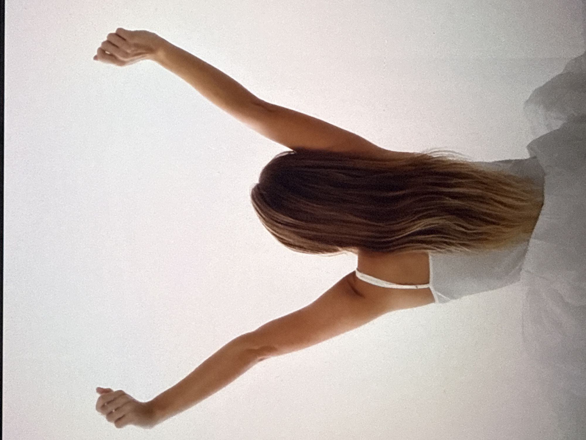 A woman with long, wavy brown hair, her back to the camera, is stretching with her arms raised above her head. She is wearing a white slip. The background is a bright, overcast sky, suggesting an indoor setting with diffused light.