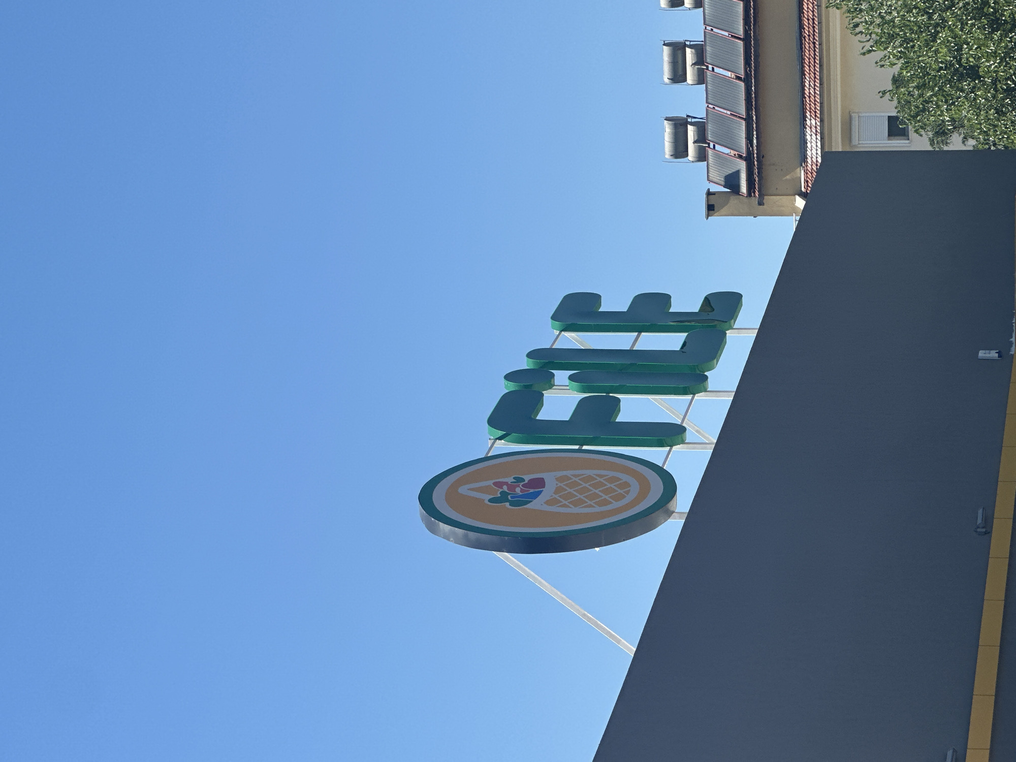 The image presents an outdoor scene dominated by a clear, bright blue sky on a sunny day. In the left-hand side, a prominent sign is visible, featuring large, green three-dimensional letters that spell out "LİFE". Below these letters, a circular logo is attached, depicting a yellow background with a green outer rim. Inside the circle, a design resembling a woven basket or waffle cone is filled with colorful produce, including red and green fruits and some blue elements. The sign is supported by visible white or light-colored metal structures.
In the lower portion of the image, the dark grey facade of a modern building or structure is visible, with a distinct yellow vertical stripe running along one edge. Behind this structure and to the right, another building stands with cream-colored walls and a reddish-brown tiled roof. Multiple solar water heaters, appearing as cylindrical tanks, are mounted on the roof, alongside several solar panels. A single window with a white blind is visible on the cream wall, and lush green foliage from a tree frames the top right corner of this building. The bright light and clear sky suggest it is midday or early afternoon. Given the location context of Fethiye, Turkey, the presence of solar water heaters and the style of the sign are consistent with common architectural and commercial elements in the region.