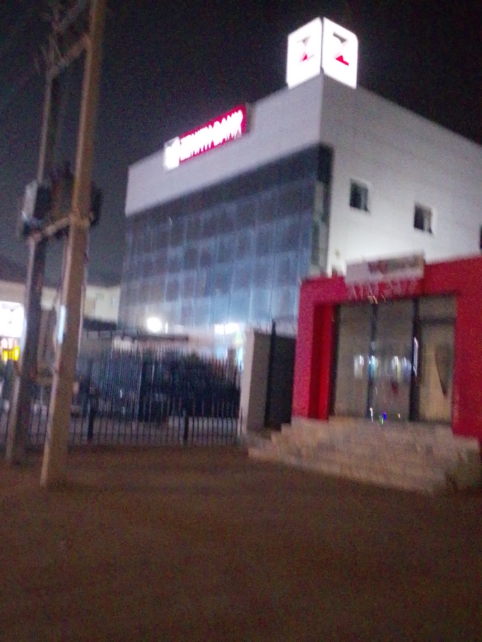 This nighttime image depicts the exterior of a Zenith Bank branch located in Gandu Albasa, Nigeria. The central subject is a multi-story building featuring a white upper facade and a lower section clad with extensive dark-tinted glass panels. A prominent, illuminated red "ZENITH BANK" sign is centrally positioned on the white facade, while an illuminated white "Z" logo marks the rooftop. To the right, a red-framed entry alcove with glass doors leads into the building. The ground level in front of the building is secured by a dark metal fence with vertical bars, behind which dark foliage is visible. The immediate foreground consists of dark, unpaved ground. On the left side of the frame, a utility pole with multiple cross arms and electrical components stands tall. No individuals are visible, and no actions or interactions are depicted. Artificial lighting from the bank's signage and interior illuminates the scene against a dark night sky.