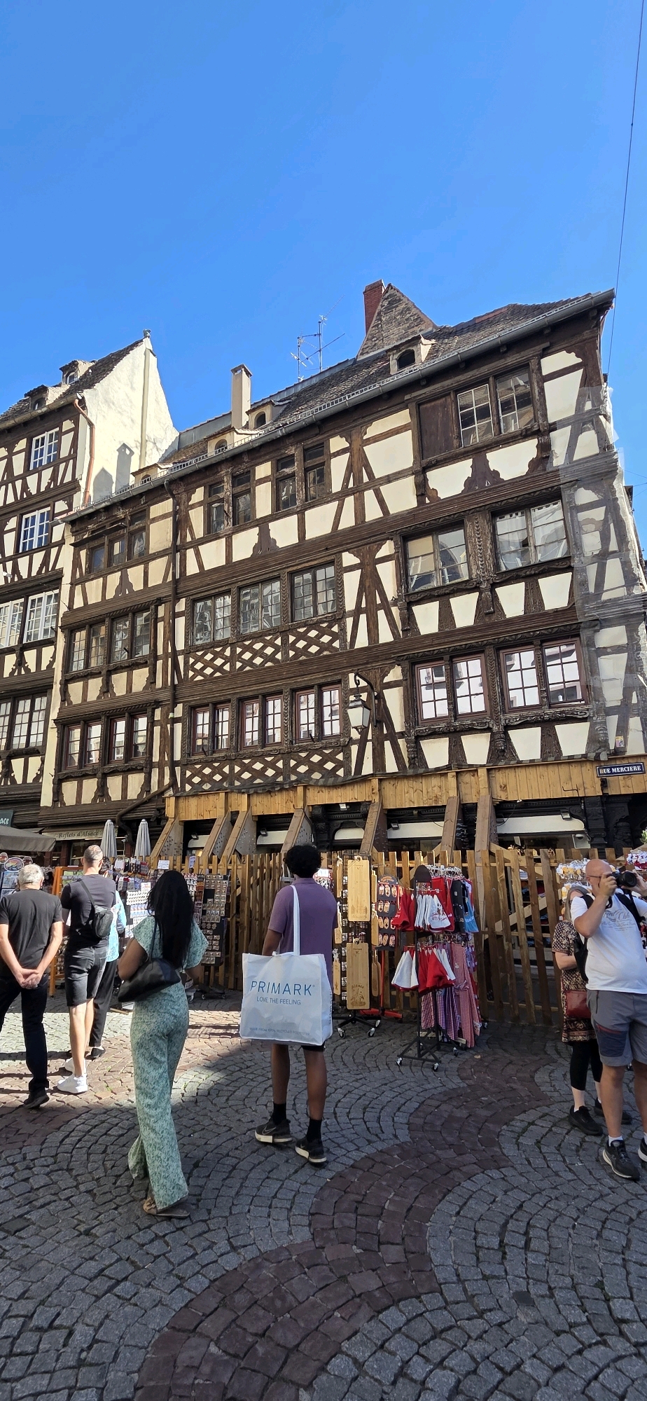 The image shows a street scene in Strasbourg, France, with a focus on the architecture of the buildings. The buildings are characterized by their half-timbered construction, a style common in Alsace. The image captures a view of several historic buildings with their distinctive dark wooden beams and lighter infill. In the foreground, several people are visible, some walking and others browsing stalls that appear to be set up in front of the buildings. One person is carrying a white shopping bag with the "Primark" logo. The ground is paved with cobblestones. The sky is clear and blue, indicating good weather. The overall scene depicts a lively urban environment in a historic district.
