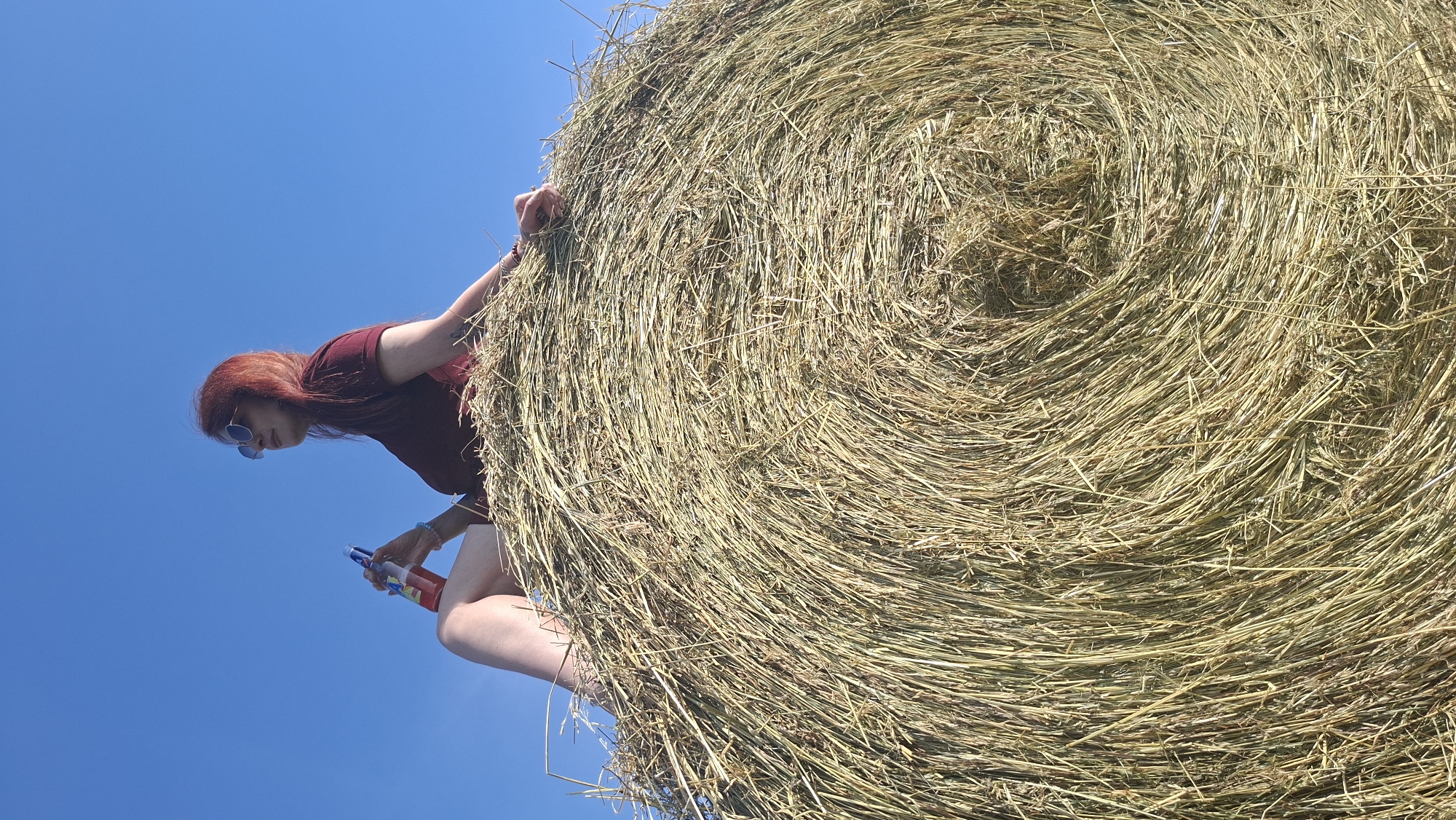 A woman with red hair sits on top of a large, round hay bale, holding a bottle of red liquid. She is wearing a maroon t-shirt and sunglasses. The hay bale is positioned in an outdoor setting with a clear blue sky as the backdrop. The image appears to be taken from a low angle, looking up at the woman and the hay bale. The location is identified as Забалоцкі сельскі Савет, Belarus.
