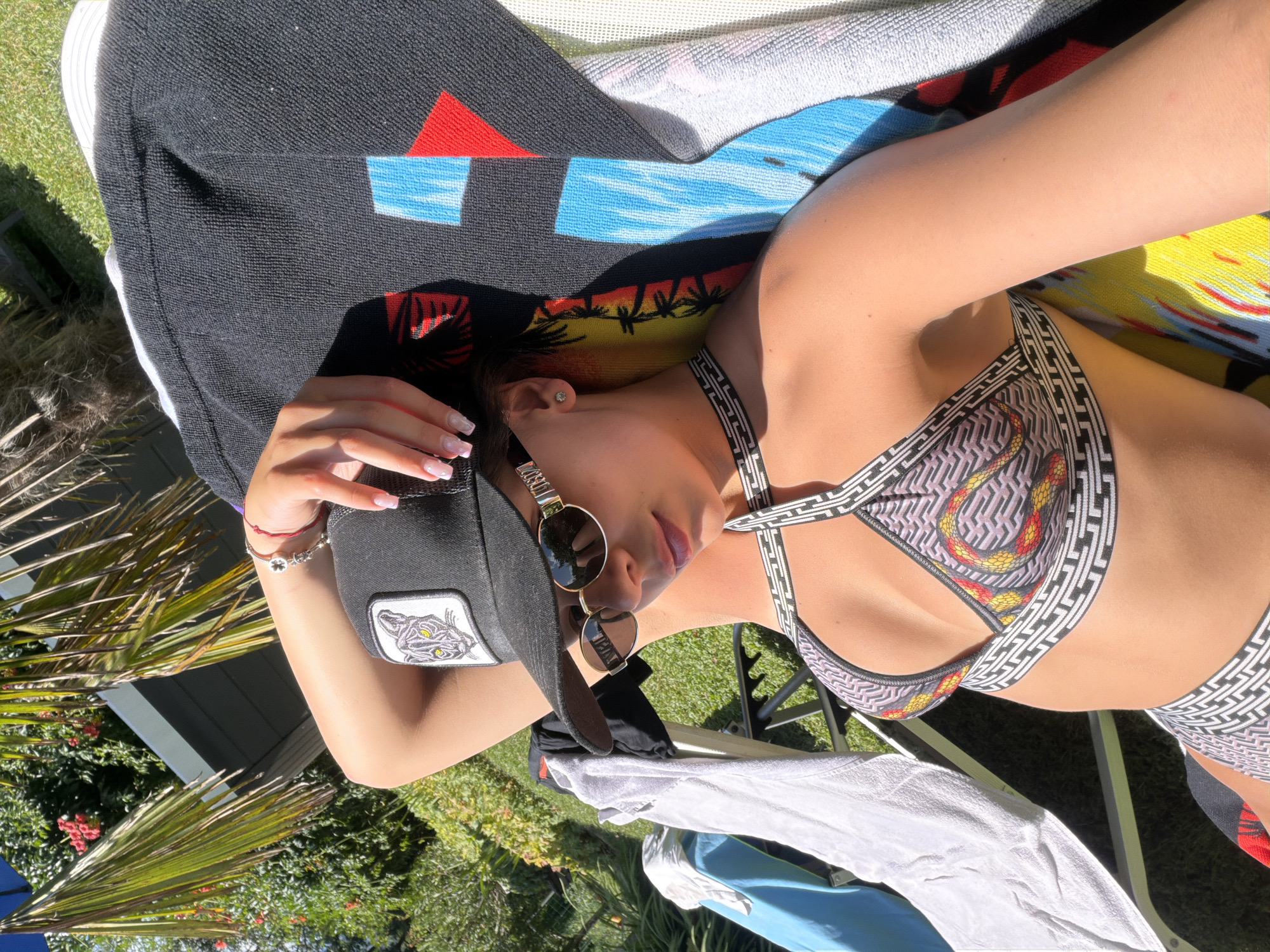 A young woman with fair skin and dark hair is lying on a lounge chair outdoors, possibly in Chavei, Bulgaria, basking in the sun. She is wearing a black baseball cap with a tiger graphic on the front, sunglasses with gold frames, and a patterned bikini. Her right arm is raised above her head, resting on the cap, and her left hand is on her hip. A patterned towel is draped over her body, and other towels and clothing are visible on the chair. The background features lush green foliage and a hint of a building. The overall scene suggests a relaxed, sunny day.