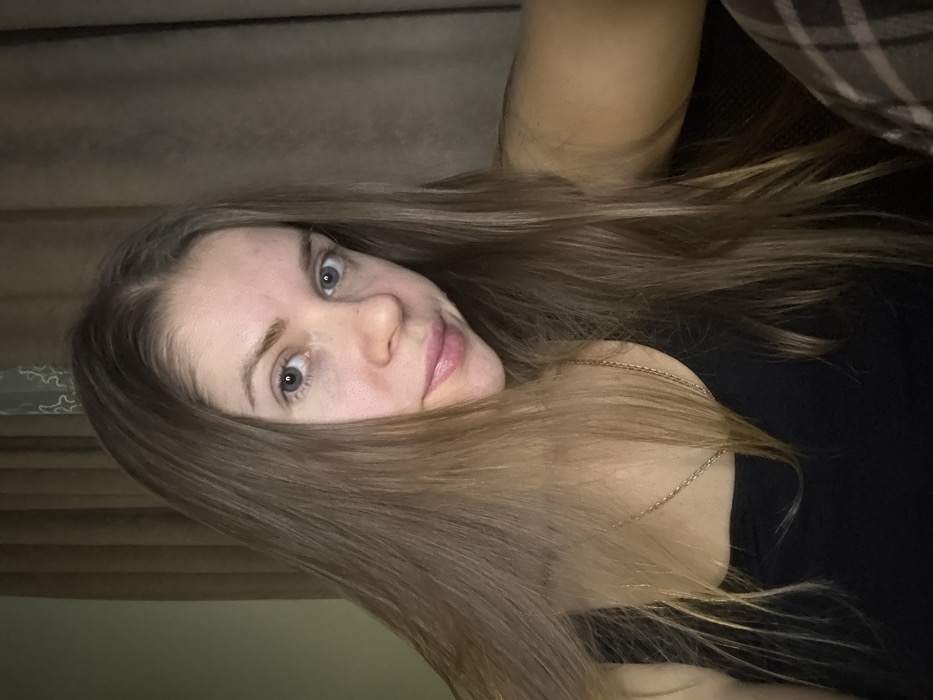 A woman with long, light brown hair partially covers her face in this selfie. Her blue eyes are prominent, and she has fair skin. She wears a black top and a delicate gold necklace. A textured curtain is visible behind her, while a portion of a plaid garment appears in the upper right corner. The image is rotated slightly.