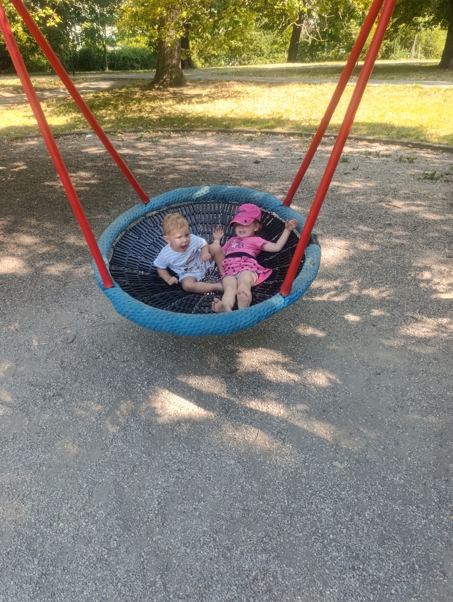 This is a photograph of two young children on a large, circular swing set in a park. The swing has a blue rope border and a black net seating area, suspended by red metal poles. On the left side of the swing, a young boy with blonde hair and a light blue striped t-shirt is sitting with his mouth open, appearing to be shouting or laughing. On the right, a young girl with blonde hair, wearing a pink t-shirt, pink shorts with white polka dots, and a pink baseball cap, is lying down. She is smiling and looking towards the left. The ground beneath the swing is covered in small, grey gravel, with patches of dried grass and tree shadows visible. In the background, mature trees with green foliage line a pathway, suggesting a sunny day in a park setting. The location is identified as the city of Olomouc, Czechia.