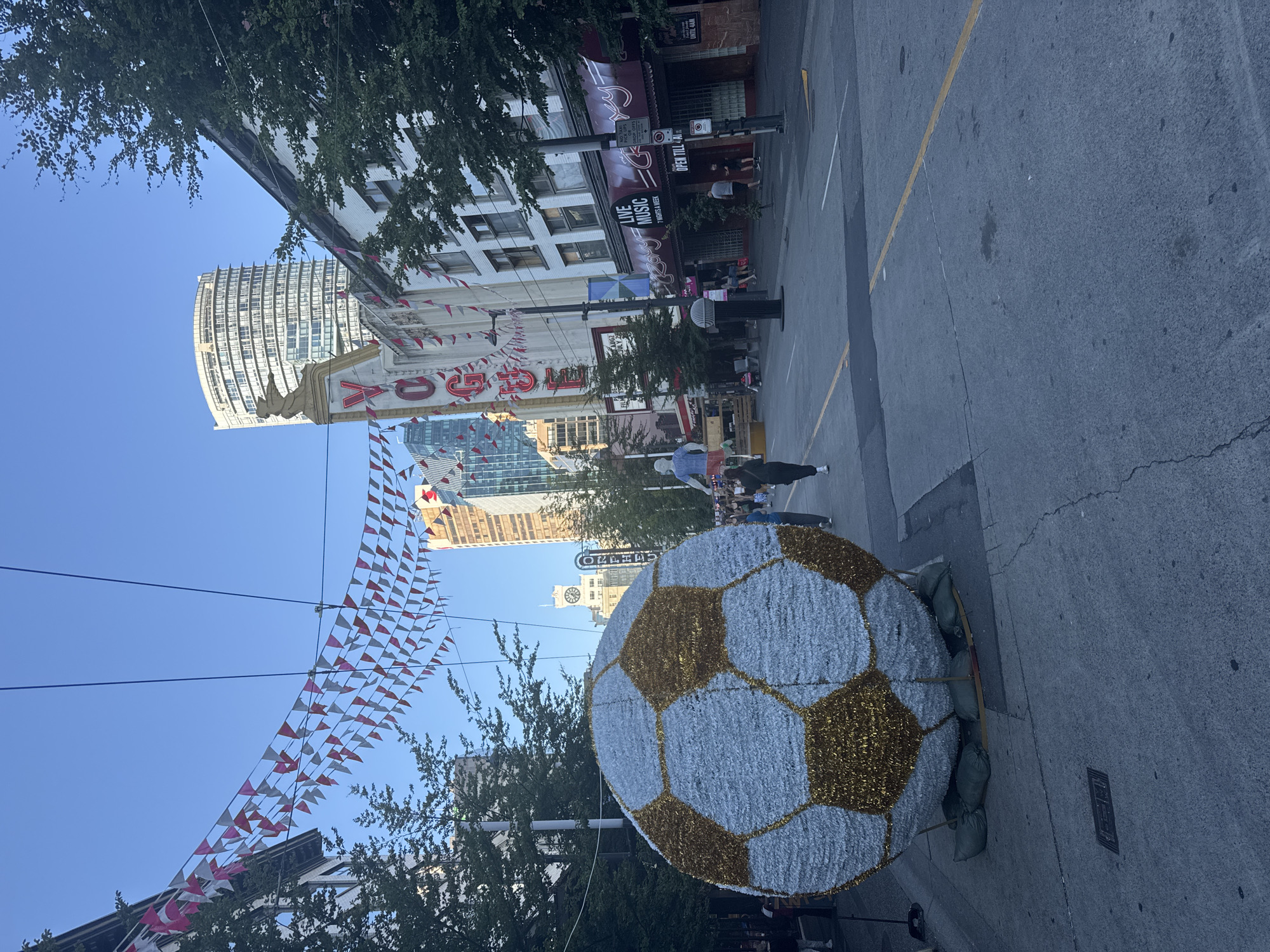 The image depicts a street scene in Vancouver, Canada, likely in the downtown core, possibly Gastown or a similar historic area given the architecture and signage. In the foreground, a very large, decorative soccer ball made of white and gold tinsel-like material dominates the left side of the frame. The soccer ball is secured to the ground with sandbags.
The street is paved with asphalt and marked with yellow and white lines. Overhead, strings of red and white triangular pennant banners are suspended across the street, suggesting a festive occasion or event.
On the right side of the street, a white building with a sign reading "VOGUE" is visible, with a stylized figure atop it. Further down, the facade of a building with a sign that reads "Roxy" and "LIVE MUSIC" is present, with patrons visible entering or exiting. Trees line the sidewalks.
In the background, modern high-rise buildings of glass and concrete contrast with the older architecture. The sky is clear and blue, indicating fair weather. A few people are walking on the sidewalk and across the street, appearing to be going about their day. The overall atmosphere suggests an urban environment during daylight hours, possibly late afternoon due to the quality of the light.