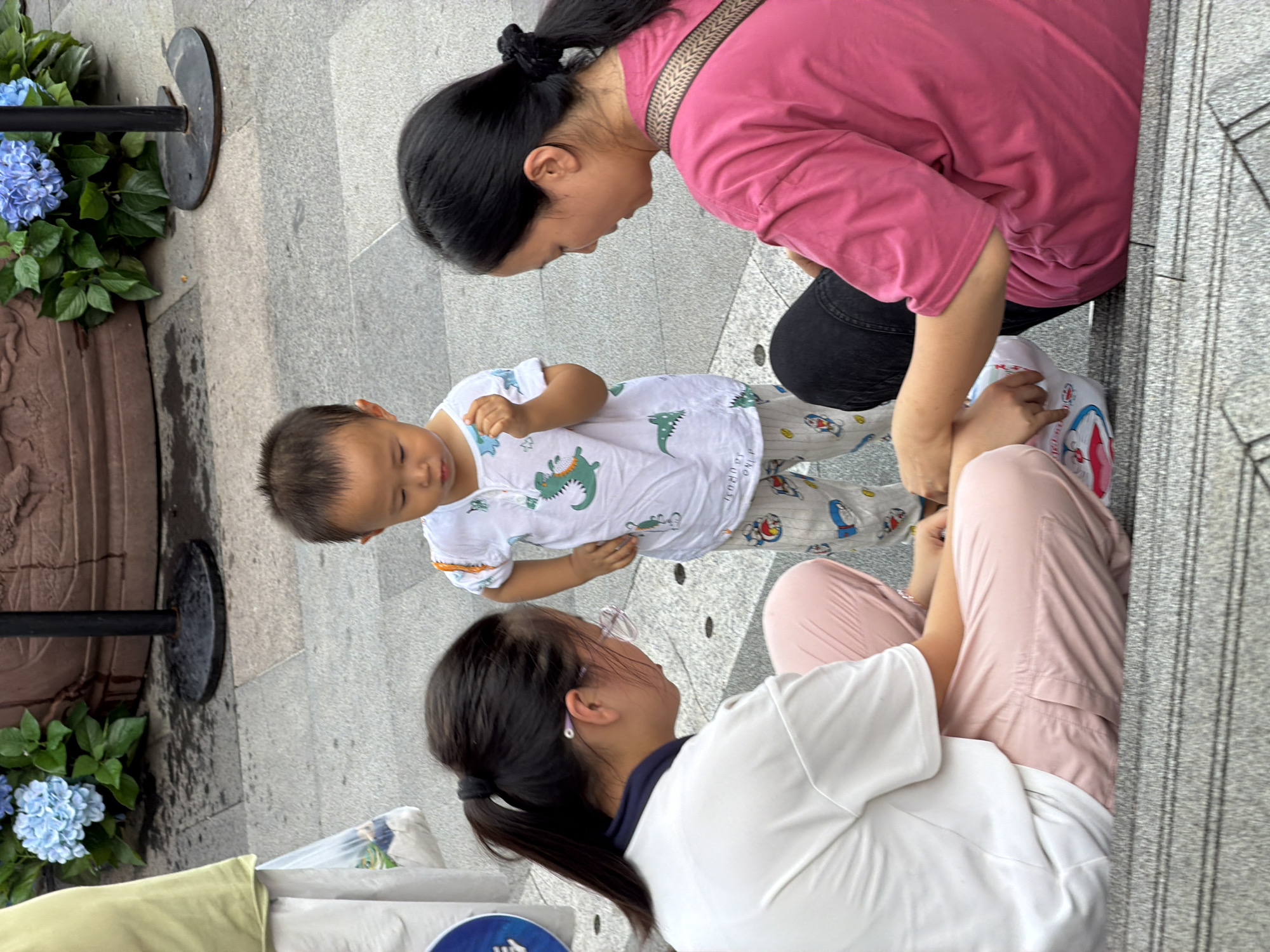 The image shows a scene in Nan'an District, China. Three people are present: an adult woman wearing a pink t-shirt and black pants, an adult woman wearing a white t-shirt and pink pants with glasses, and a young child wearing a white t-shirt with dinosaur prints and patterned pants. The two adult women are sitting on steps, and the child is standing between them. The woman in pink is holding a white object with red and blue designs. The woman in white is looking down, and the child is also looking down. The environment appears to be an outdoor public space, with a paved ground and some potted plants with blue flowers visible in the background. The lighting suggests it is daytime.
