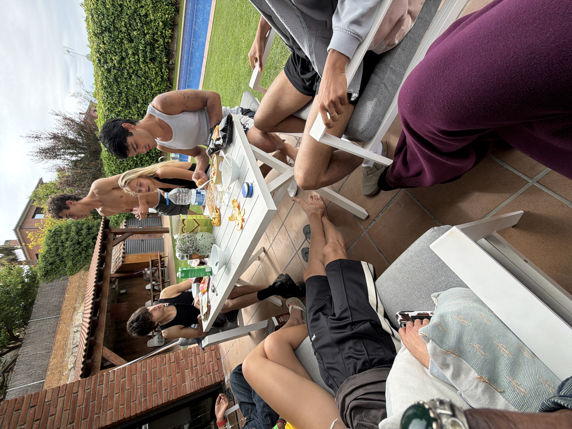 A group of people are gathered around a white table, potentially for a meal or snack, in what appears to be a backyard or patio setting. The location is Sant Andreu de Llavaneres, Spain.
Several individuals are seated on white chairs and a gray couch, with some showing bare legs and feet. There is a person wearing a gray tank top and shorts, a person wearing black shorts and sneakers, a person with bare feet, and another person with a gray hoodie. A person in a black tank top is reaching under the table, and others are focused on the food or items on the table.
The white table holds a variety of items, including food, snacks, drinks, and decorative elements like a vase of flowers and a potted plant.
The setting includes a brick building with a tiled roof, a well-maintained green lawn, and a swimming pool. The sky overhead is partly cloudy.
