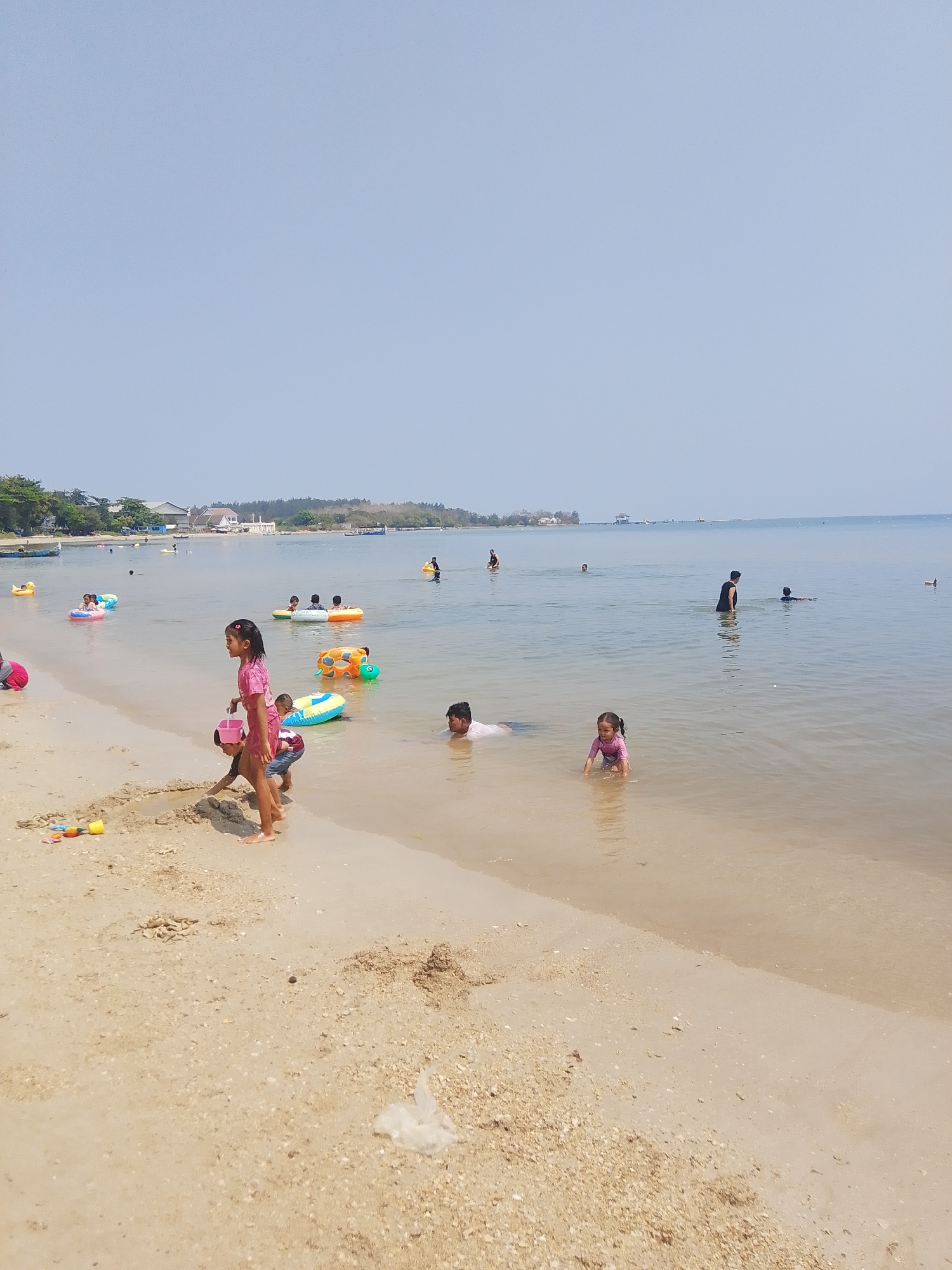 The image depicts a crowded beach scene at the coastal city of Pejagan, Indonesia, under a clear, pale blue sky. The shallow, calm water extends to the horizon, where a hazy landmass is visible. Numerous people, predominantly children, are engaged in various water activities. Several children are playing in the sand near the water's edge, equipped with beach toys. Others are in the water, some using inflatable rings, including one shaped like a turtle, and others simply wading or swimming. Adults are also present, supervising and participating in the activities. The beach itself is sandy and appears to be a popular recreational area.