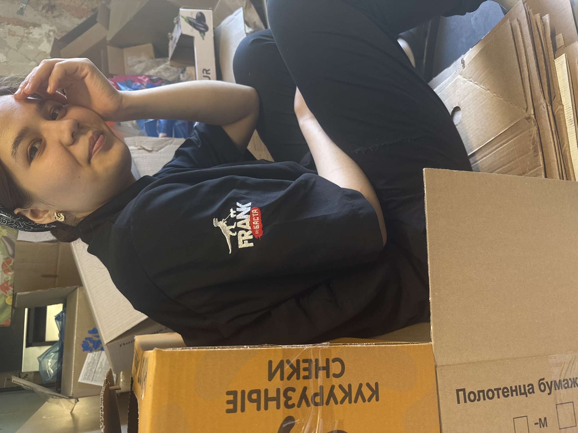 The media file shows a young woman lying down in a cluttered environment, surrounded by cardboard boxes. She is wearing a black t-shirt with a logo on the left chest that appears to read "FRANK" with some other text. She has her left hand on her forehead, and her right arm is bent, resting on her leg. She is looking towards the camera with a neutral expression. She is wearing black pants and a black and white patterned bandana or hair covering.
The background is filled with various cardboard boxes, some stacked, others open, suggesting a storage area or a place where items are being unpacked or packed. Some boxes have text printed on them in Russian. One prominent box in the foreground has the text "КУКУРУЗНЫЕ СНЕКИ" (corn snacks) printed in large letters. Another box to the right has the text "Полотенца бумажные" (paper towels) and a size indication "-M". There are also glimpses of other items, including blue plastic bags.
The lighting appears to be artificial, casting a soft glow on the scene. The overall impression is one of a casual, perhaps messy, setting, with the woman seemingly at ease amidst the boxes. The time of day and weather are not discernible from the image. The location is not specified but appears to be indoors.