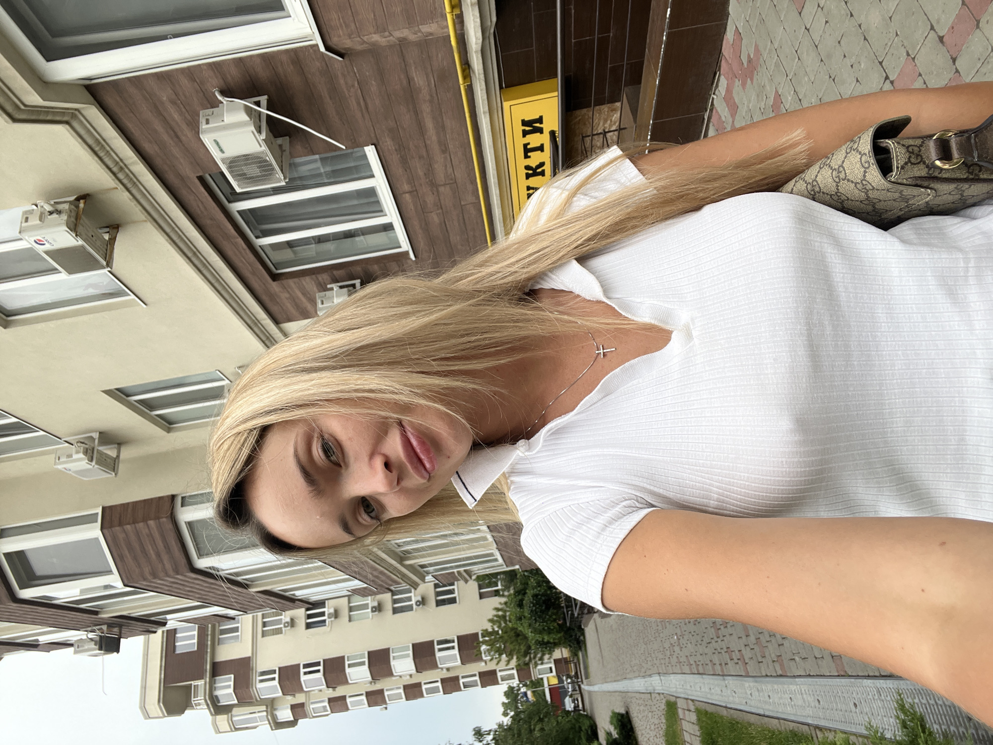 A medium shot, eye-level, selfie taken outdoors in Kryzhanivka, Ukraine. A blonde woman with fair skin and light blue eyes is in the foreground, looking at the camera with her lips slightly parted. She is wearing a white ribbed polo shirt and a silver cross necklace. To her right, she holds a beige and brown Gucci shoulder bag. Behind her, there are several modern, multi-story buildings with light yellow and brown facades, featuring numerous windows and external air conditioning units. A paved walkway with a grass border is visible to the left, and a yellow sign with dark lettering is mounted on one of the buildings. The sky is overcast.