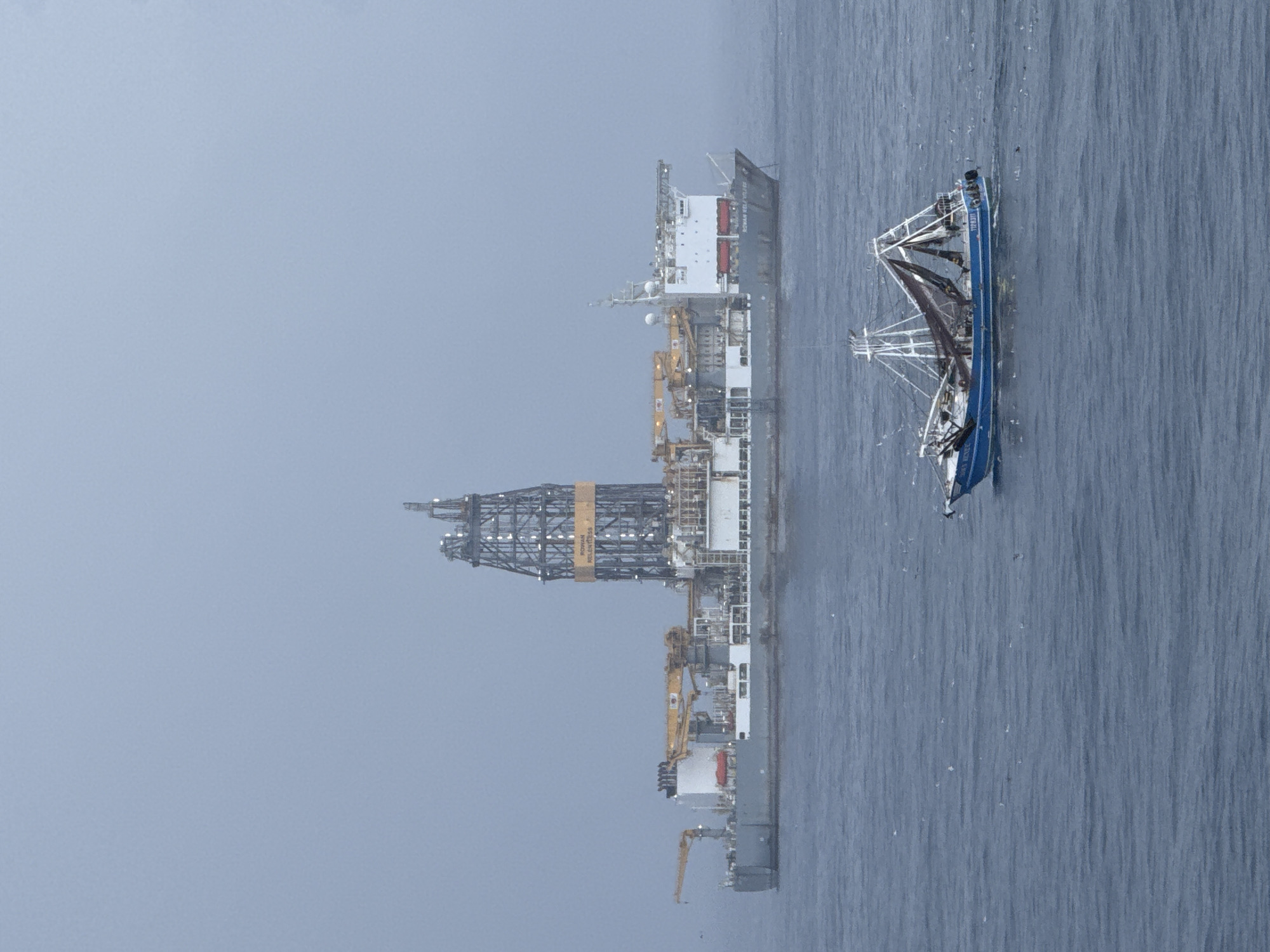 The image shows two vessels on the water under a cloudy sky. On the left is a large semi-submersible oil drilling rig named "ROWAN RELENTLESS". Next to it, on the right, is a fishing vessel. The location is Unknown, Unknown.