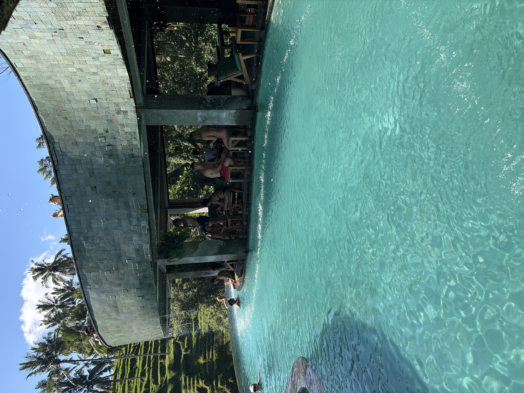 In Tegallalang, Indonesia, a curvilinear structure with a tiled exterior overlooks a turquoise swimming pool. People are visible in and around the pool and under the structure, some swimming, others seated at tables, and some standing. Lush green terraces and palm trees are seen in the background, under a bright blue sky with scattered clouds. The scene suggests a resort or cafe setting by the water.