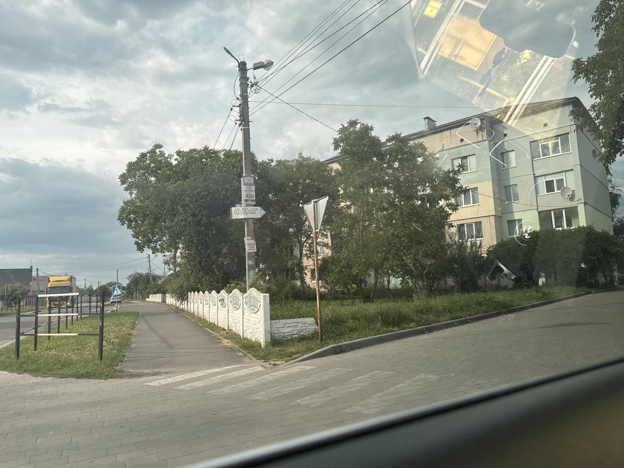 This image captures a street scene in Suprunivka, Ukraine, from inside a vehicle, indicated by the dark gray trim along the bottom edge and reflections in the upper right. The sky is heavily overcast with dense cloud cover.
In the immediate foreground, a light grey paver sidewalk transitions into a paved crosswalk marked with white stripes. To the left of the crosswalk, a small strip of grass with scattered weeds separates the pedestrian area from a section of black metal barriers with horizontal yellow/white stripes, extending towards the left.
The midground features a paved road extending into the distance on the left, with a large yellow truck partially visible further down. Parallel to this road, on the right, runs another paved path or sidewalk. This path is bordered on its right by a low, white decorative concrete fence with arched sections. A tall, weathered concrete utility pole stands prominently near the center-right, supporting multiple overhead power lines and a streetlight fixture. Attached to the pole are several signs: a purple rectangular sign at the top with Cyrillic text including "frugat," a white rectangular sign below it displaying "850" and additional Cyrillic text, and a white arrow sign pointing right, marked with the Cyrillic word "ОДЯГ" (clothing). To the right of the pole, a white triangular yield sign is partially visible. Further down the main road on the left, a blue square pedestrian crossing sign is visible.
In the background, behind dense green trees and foliage, a multi-story residential building, likely an apartment block, is visible on the right. The building's exterior features sections painted in light blue/teal, light yellow, and light grey/beige. Multiple windows and at least two satellite dishes are mounted on its facade. A distinct, inverted reflection, possibly of the vehicle's interior or another structure, overlays the upper right portion of the image, including part of the building and the sky.