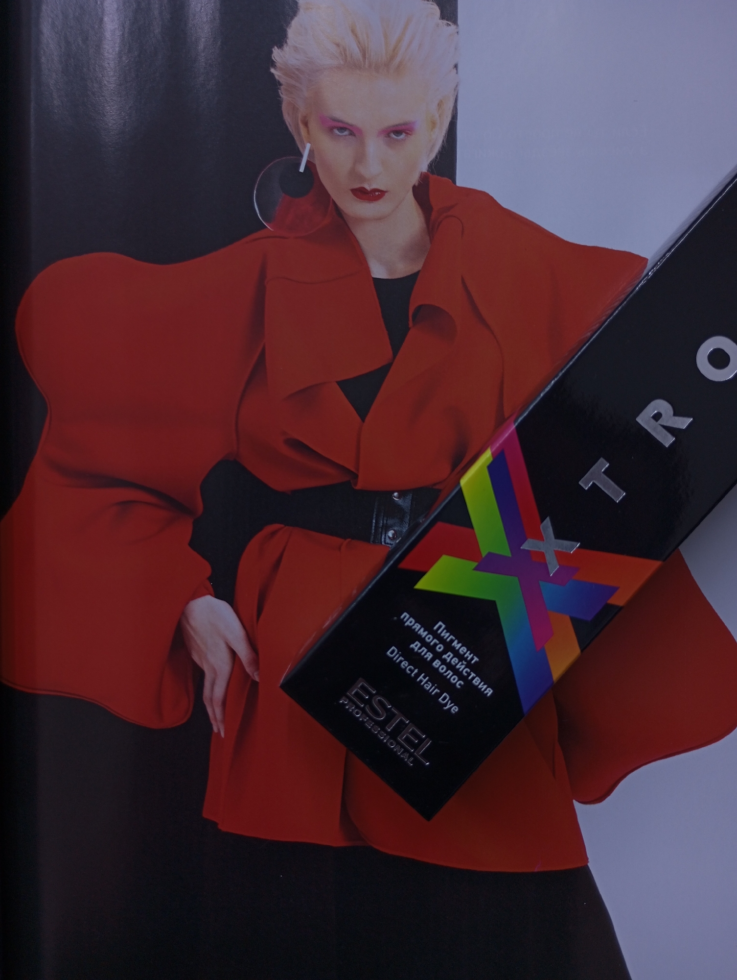The image features a fair-skinned woman with short, styled, platinum blonde hair, red lipstick and pink eyeshadow. She is wearing a black top under a belted, oversized, bright red jacket. Her pose includes an outward-facing palm. She is wearing an oversized, geometric earring on her left ear. A black box angled diagonally in the foreground partially obscures the image. The box contains the text "XTRO" in silver lettering and, below that, an abstract multi-colored pattern. Below the pattern, text in Russian reads "Пигмент прямого действия для волос," and beneath that is the English translation, "Direct Hair Dye." The box also contains the logo "ESTEL PROFESSIONAL." The background is a dark gradient, suggesting a studio or indoor setting. There is no information in the image to indicate that this image was taken in the city of Izhevsk, Russia.