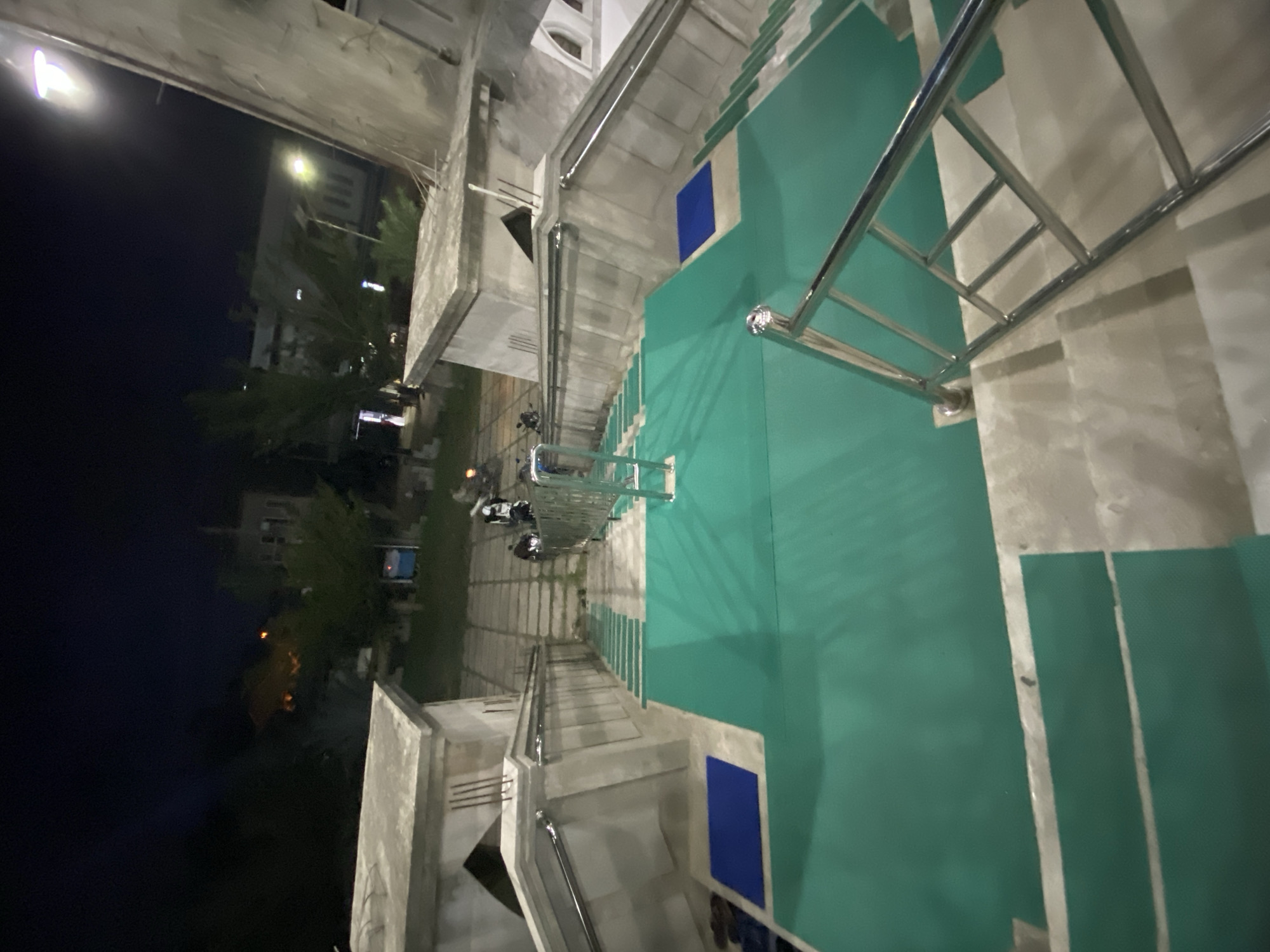 This image captures a wide-angle view of an outdoor staircase at night, likely part of a public building or religious site in Lhokseumawe, Indonesia. The staircase is illuminated by artificial lights, casting a dim glow on the surroundings. The steps are covered with a green matting, and blue mats are placed at intervals. Chrome handrails run along both sides of the stairs, providing support. In the background, several people on motorcycles are visible, suggesting a public gathering or the presence of a nearby road. The architecture of the building suggests it could be a mosque or a similar institutional structure, typical in Indonesian cities like Lhokseumawe. The overall atmosphere is quiet and serene, characteristic of nighttime in a less busy urban area.